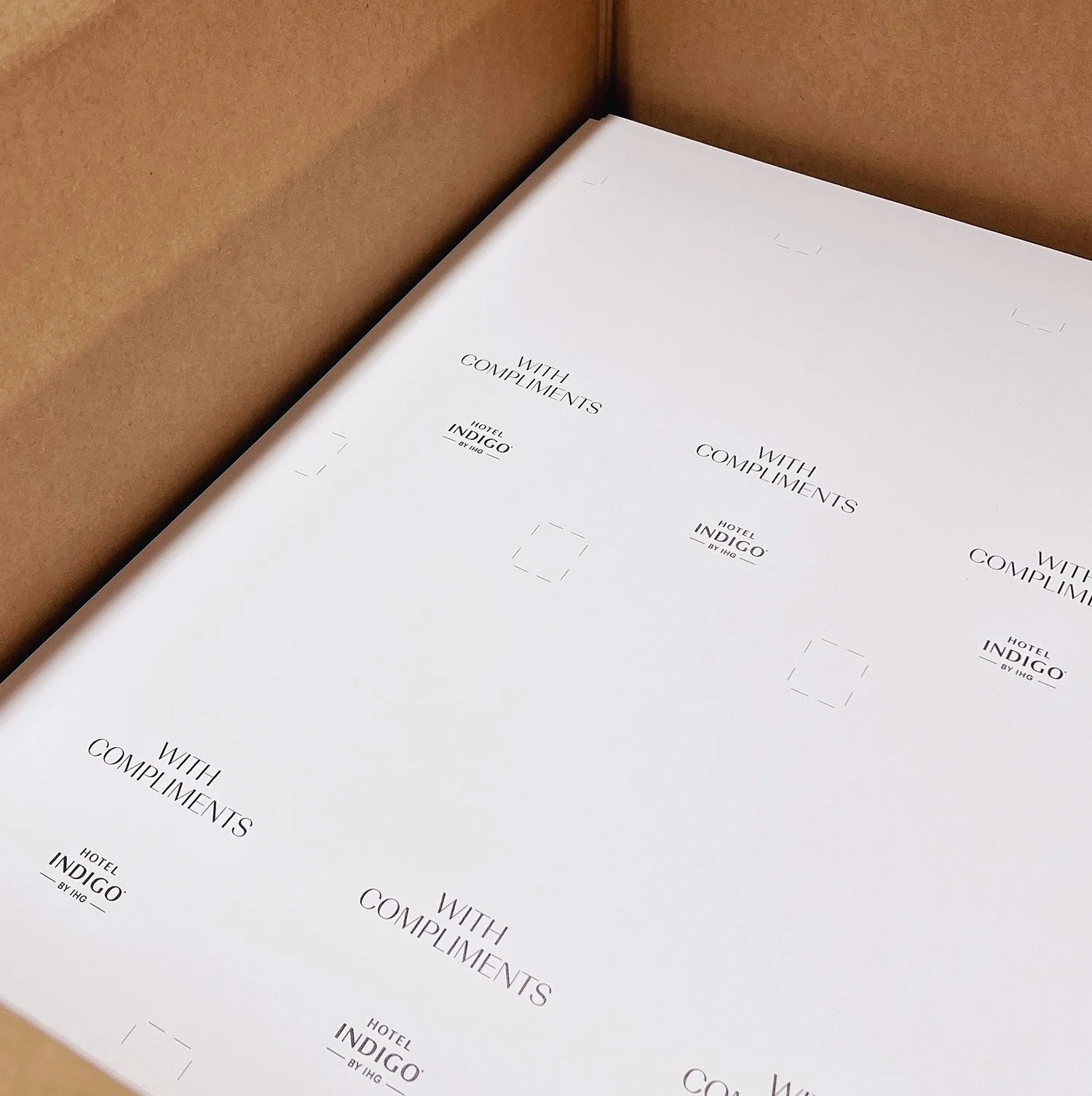 Close-up of printed paper inside a cardboard box, with repeated text 'WITH COMPLIMENTS' and 'HOTEL INDIGO BY IHG', featuring dotted registration marks.