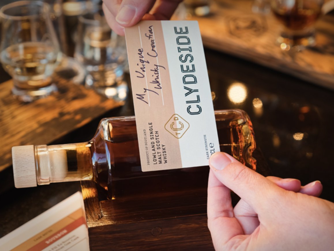 A hand holding a small bottle of whisky with a label that reads "My Unique Whisky Creation, CLYDESIDE," in front of a bar counter with glasses and a wooden surface.