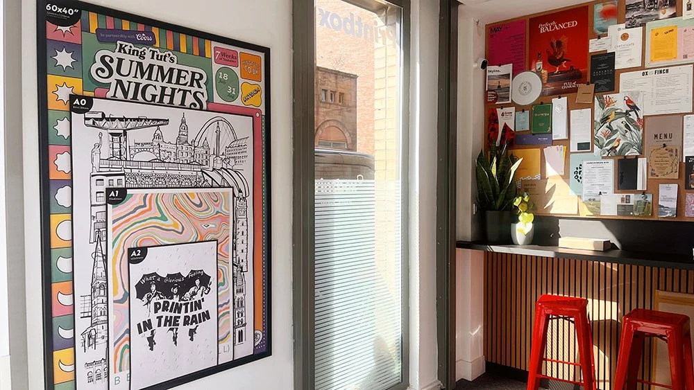Interior of a cafe or office with a colorful poster about summer nights on the wall, a corkboard with various notices and artwork, a window with blinds, a potted plant, and red stools.