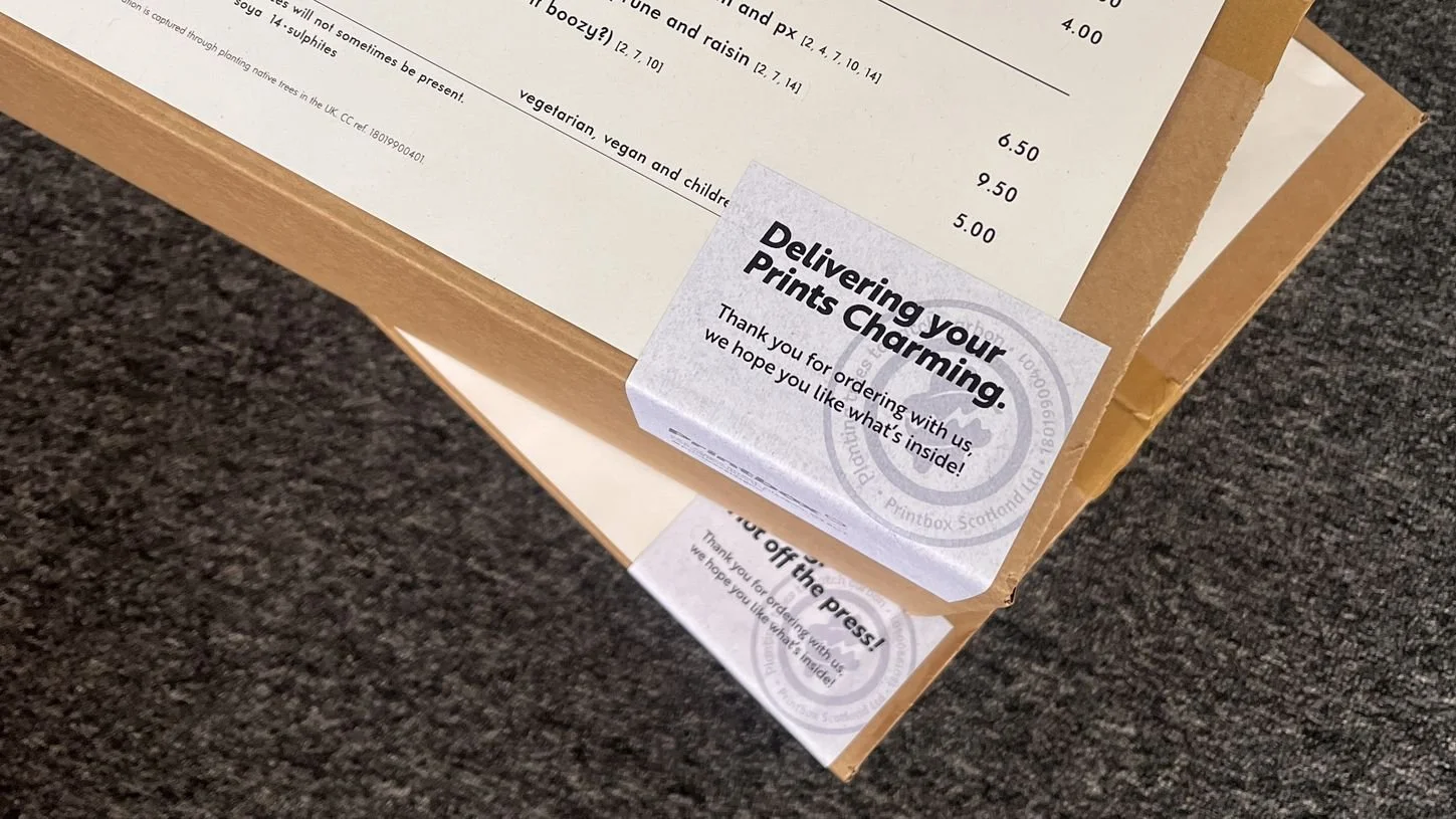 A cardboard food delivery box with a printed menu and two stickers that read 'Delivering your Prints Charming' and 'Drop off the Press!'.