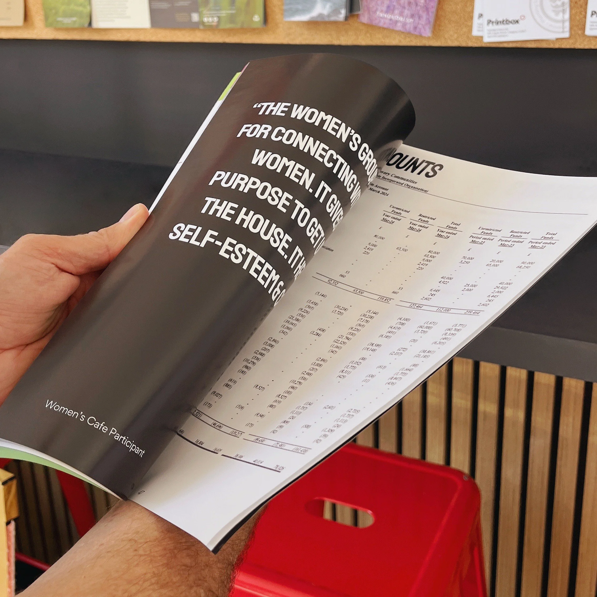 A person is holding an open magazine or booklet with a dark background page featuring white text about women. The text reads, "The women’s group for connecting women. It gives purpose to get... the house. it... self-esteem." The opposite page contains numerical data, possibly financial or statistical tables, with the surroundings including a black surface and a red plastic chair.