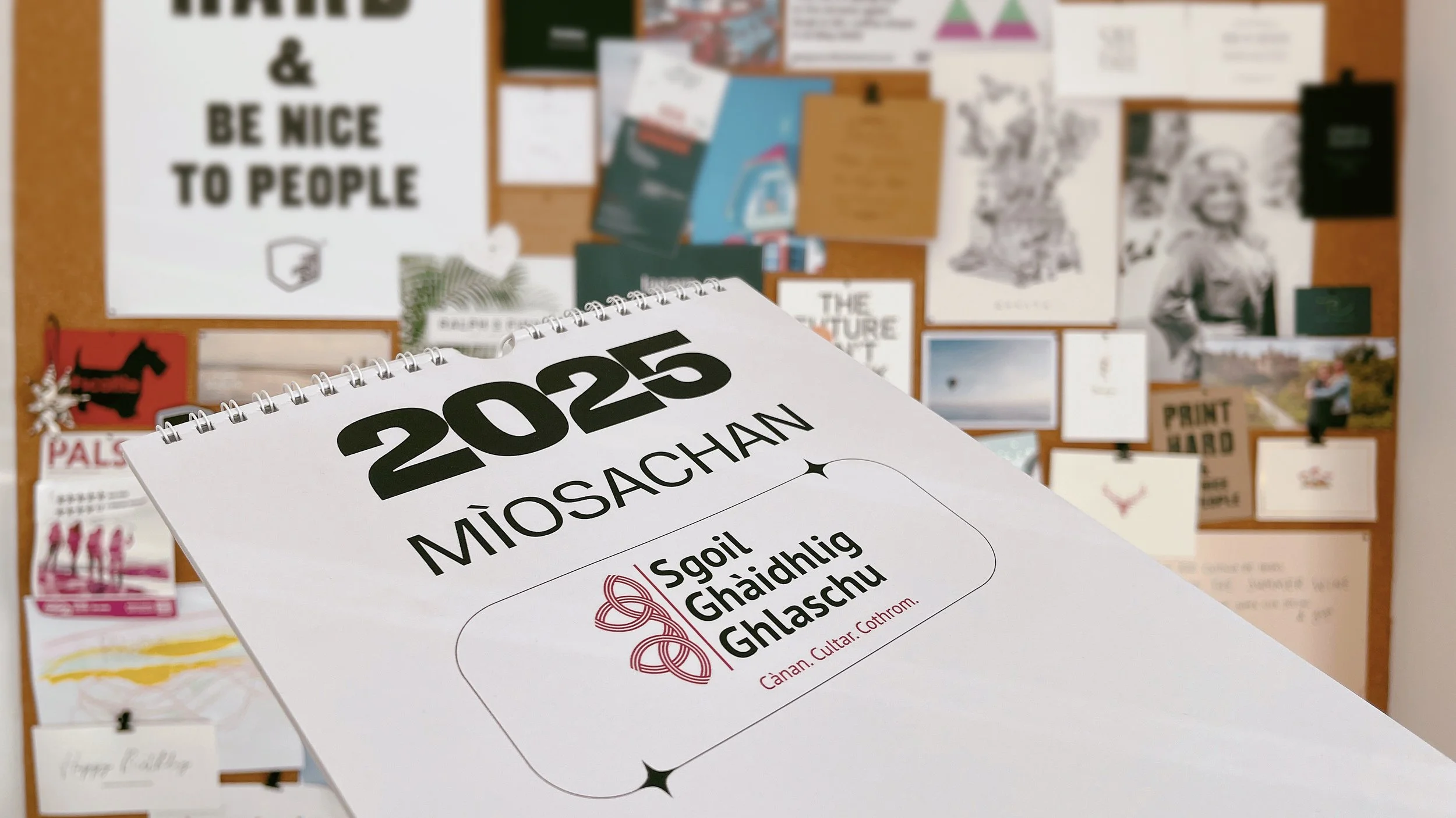 A 2025 calendar for Miòsachán featuring a logo for Sgoil Ghaidhlig Ghlaschu on a bulletin board covered with various posters and images.