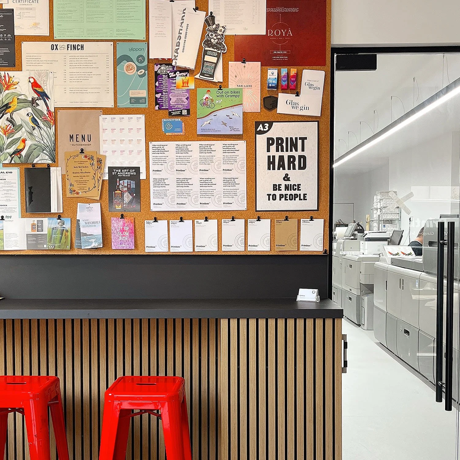 A cork bulletin board filled with various posters, flyers, and notices in a store or office, with a section of a wooden counter and two red chairs in the foreground. To the right, there is a glass wall revealing a service area with machines and equipment.