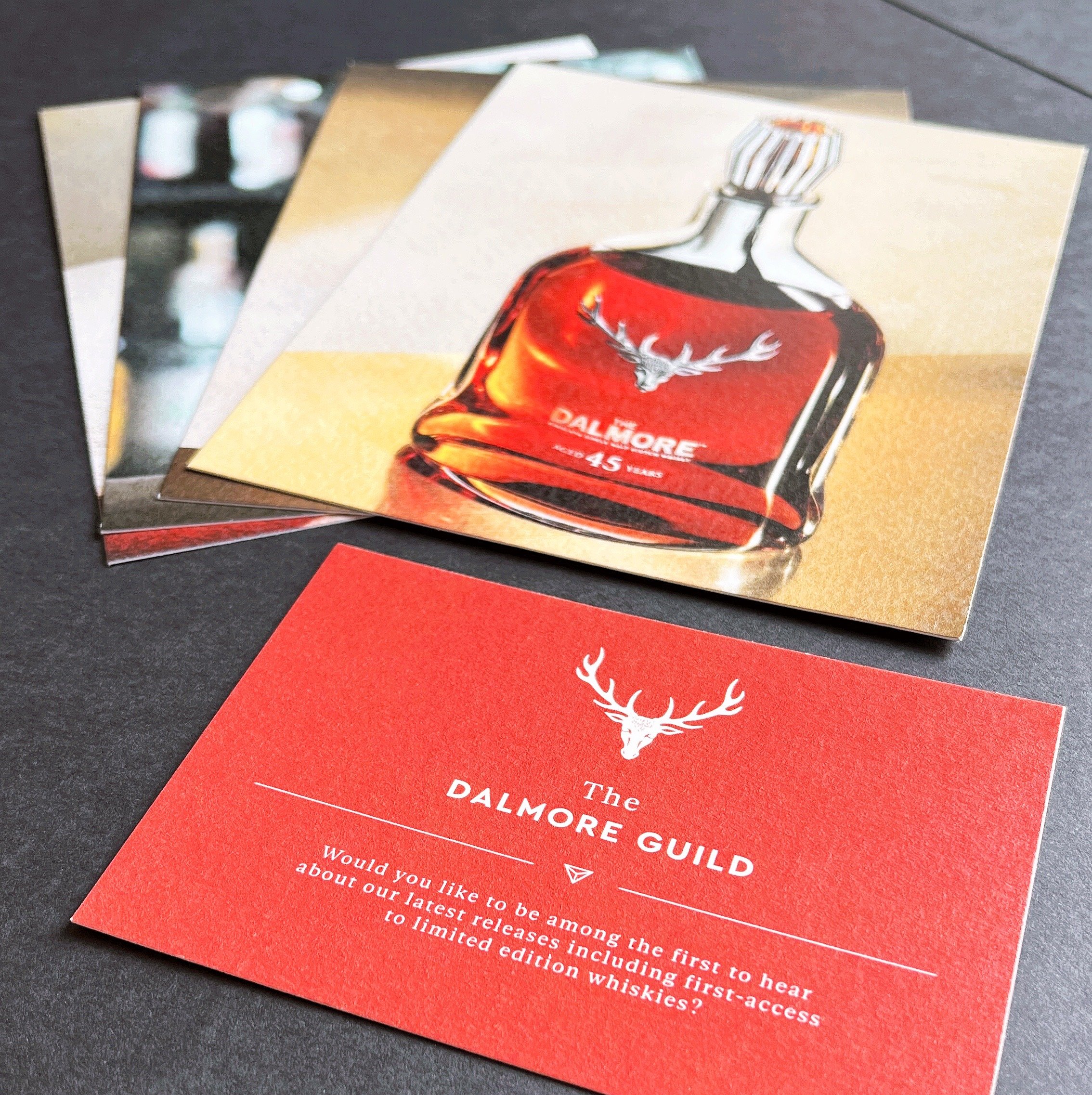 A set of promotional cards for Dalmore whisky, featuring one card with a bottle of Dalmore whisky and a red background with a white deer logo, placed on a dark surface.