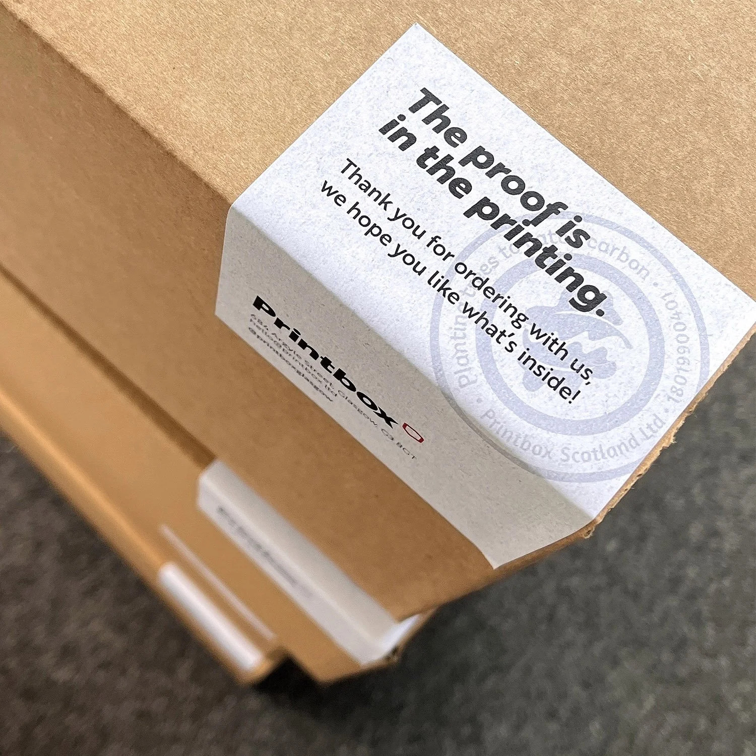 Close-up of a cardboard box with a white sticker that says, 'The proof is in the printing. Thank you for ordering with us. We hope you like what's inside!' and a Printbox logo