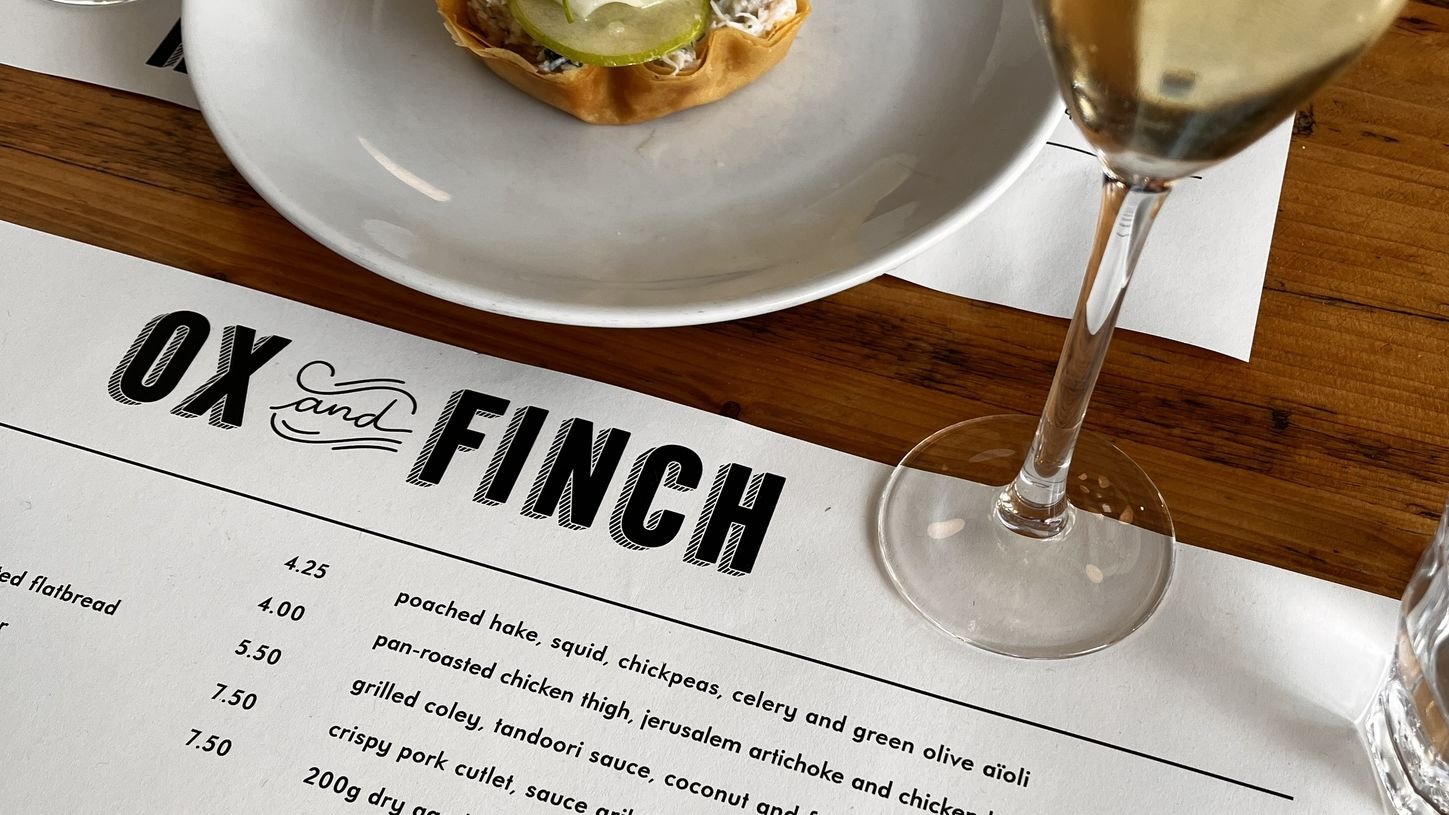 A restaurant table setting with a menu titled 'OX and FINCH', a white plate with a sandwich topped with pickles, and a glass of white wine.