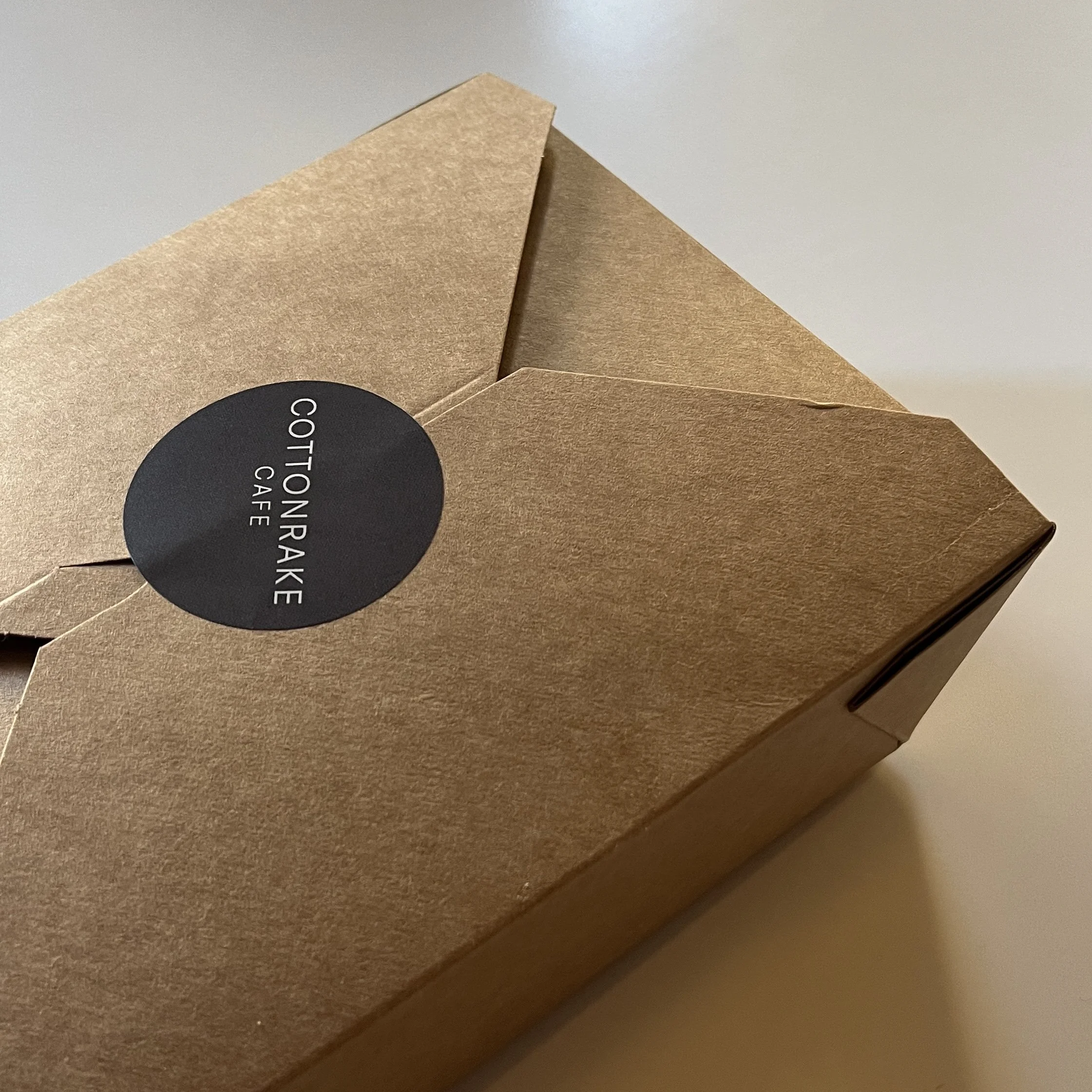 A rectangular brown paper takeout box with a black circular sticker that reads "COTTONRAKE CAFE."