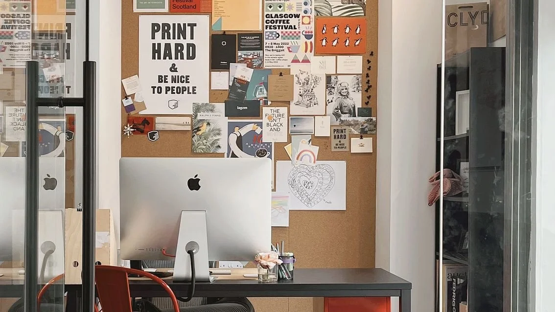 An office workspace with a corkboard filled with colorful posters and artwork, a desk with an Apple computer, and shelves with various items, all visible through glass doors.