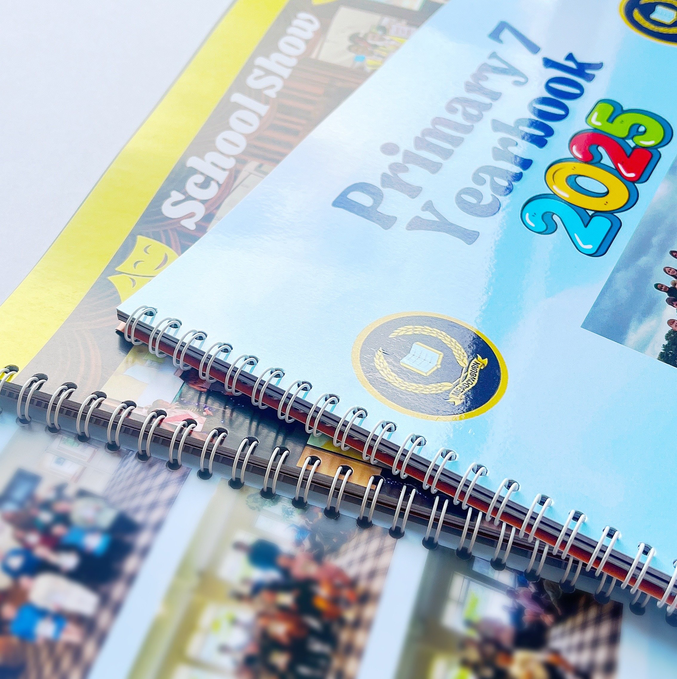 Close-up of a spiral-bound yearbook with a light blue cover titled "Princeton Yearbook 2025." The yearbook features a school emblem with a book and a wreath. Behind it, there are additional yearbooks with visible text and photographs of groups of people.