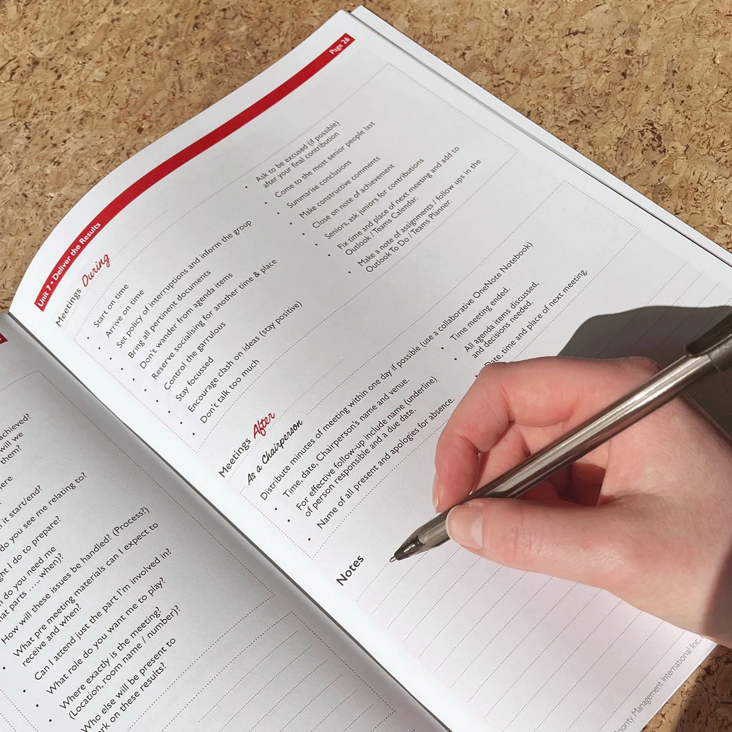 Open notebook on a wooden surface with hand writing notes with a pen, with printed text and headings about meeting procedures and a section labeled Notes.
