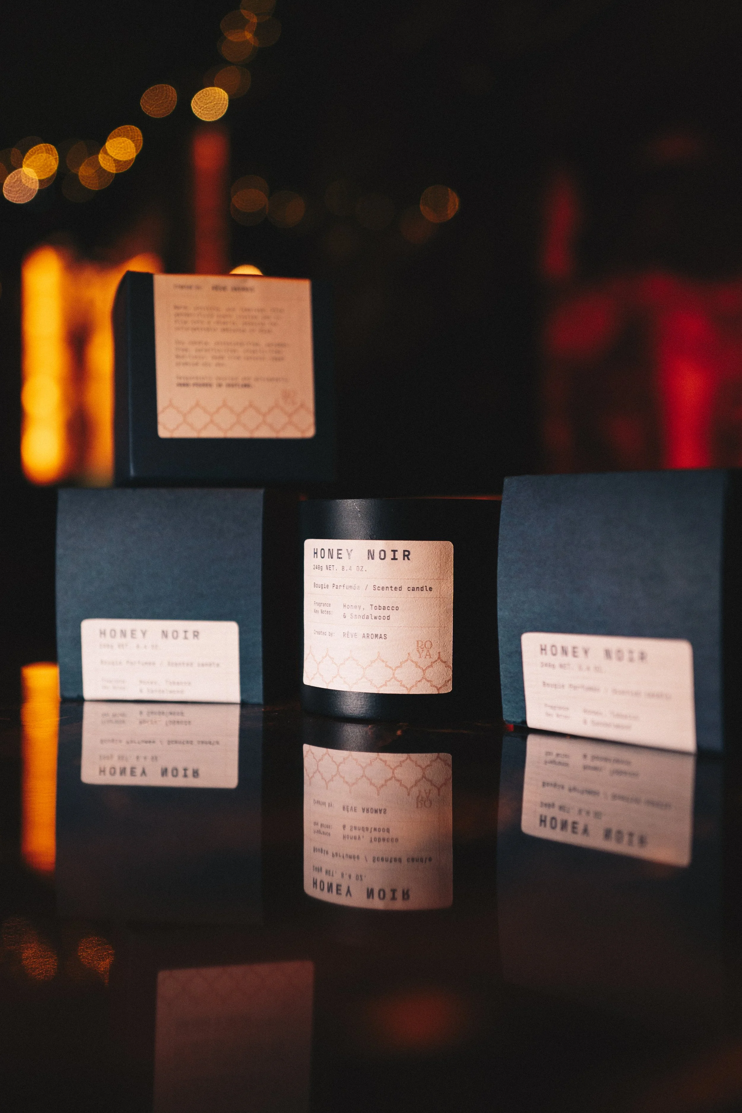Black candle labeled 'Honey Noir' with a white label, set against a dark background with bokeh lights, reflected on a glossy surface.