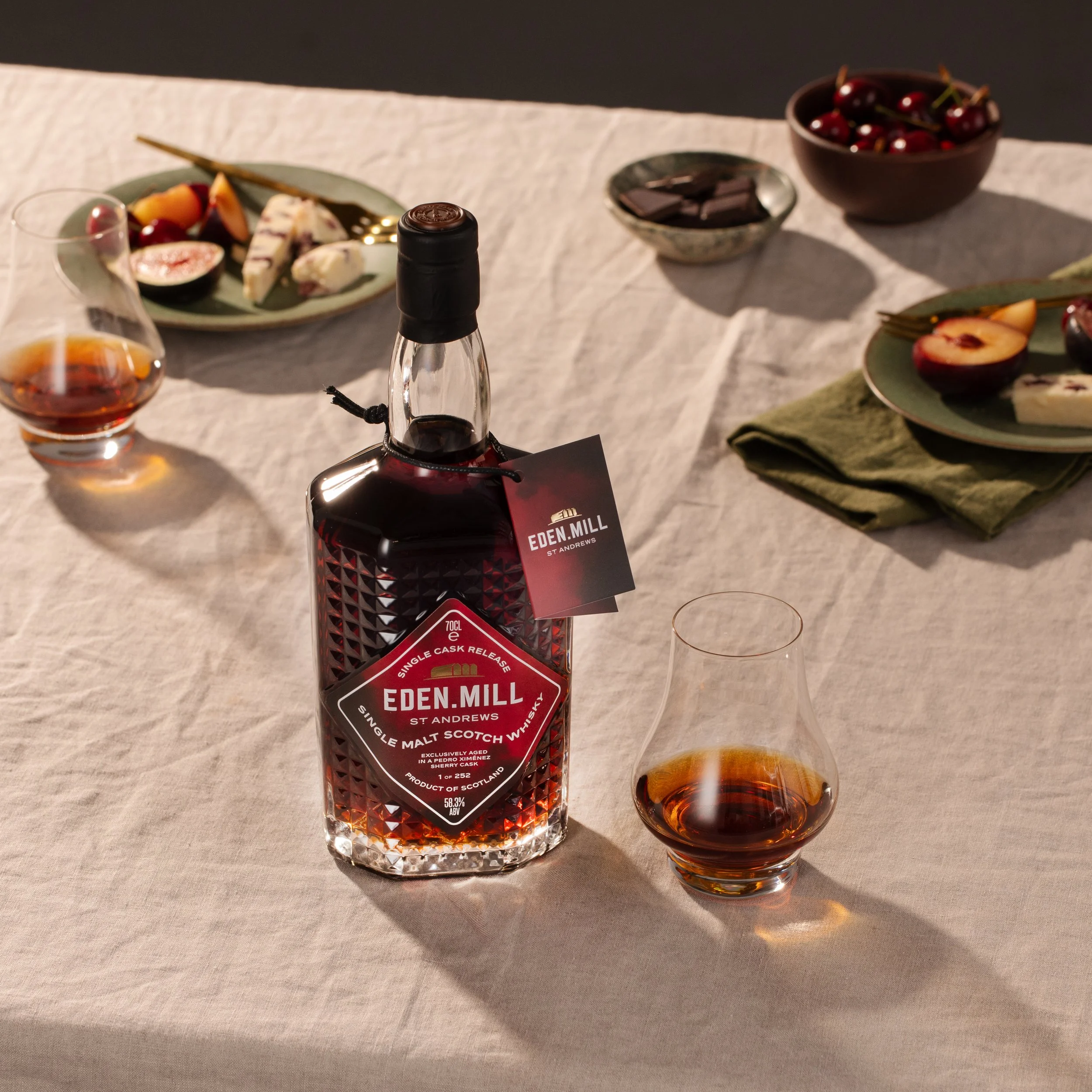Bottle of Eden Mill single malt scotch whiskey on a table with a glass of whiskey and plates of fruit and chocolates in the background.