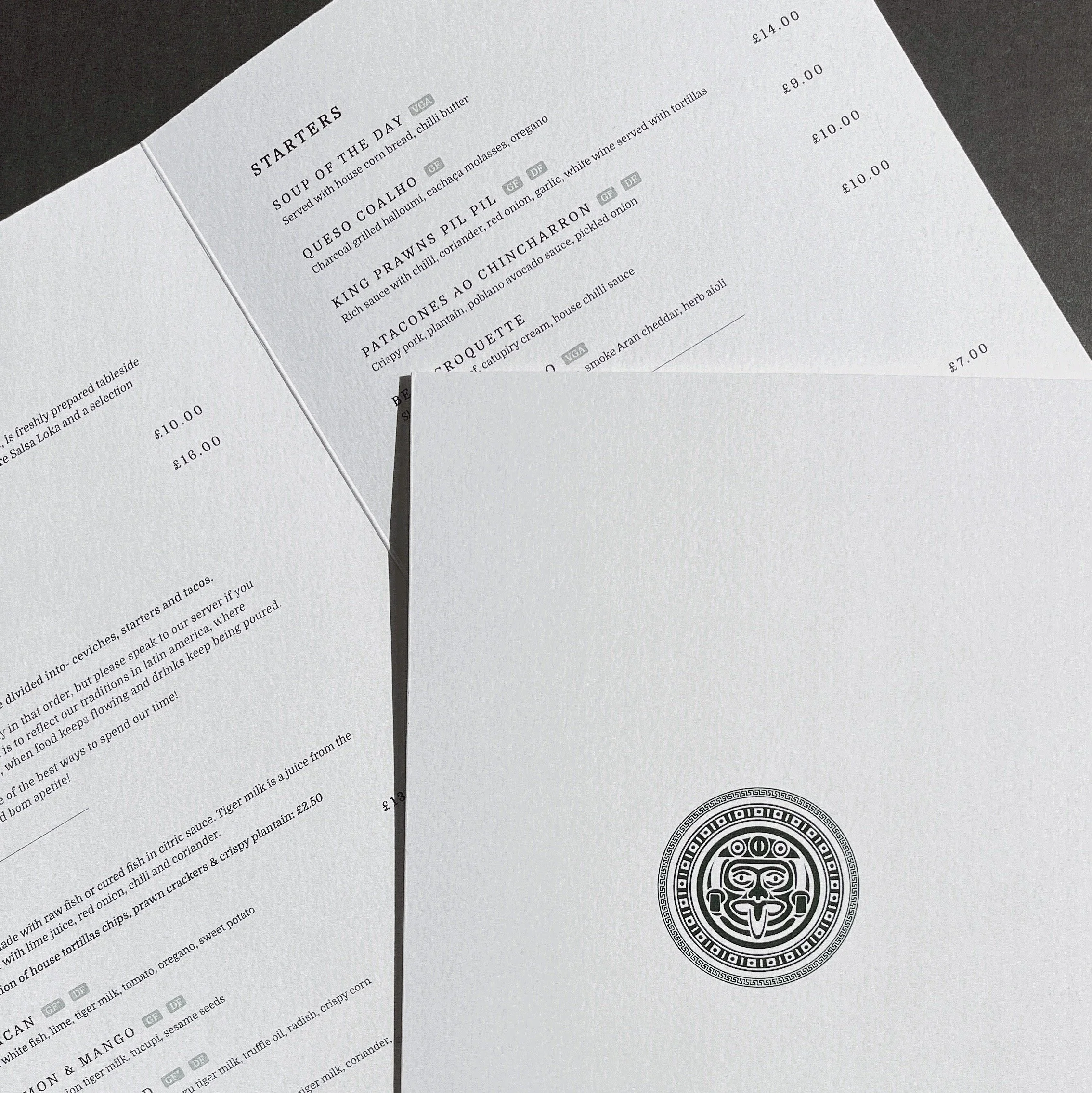 Restaurant menu pages with dishes and prices, featuring a design element or logo on a white page, placed on a dark surface.