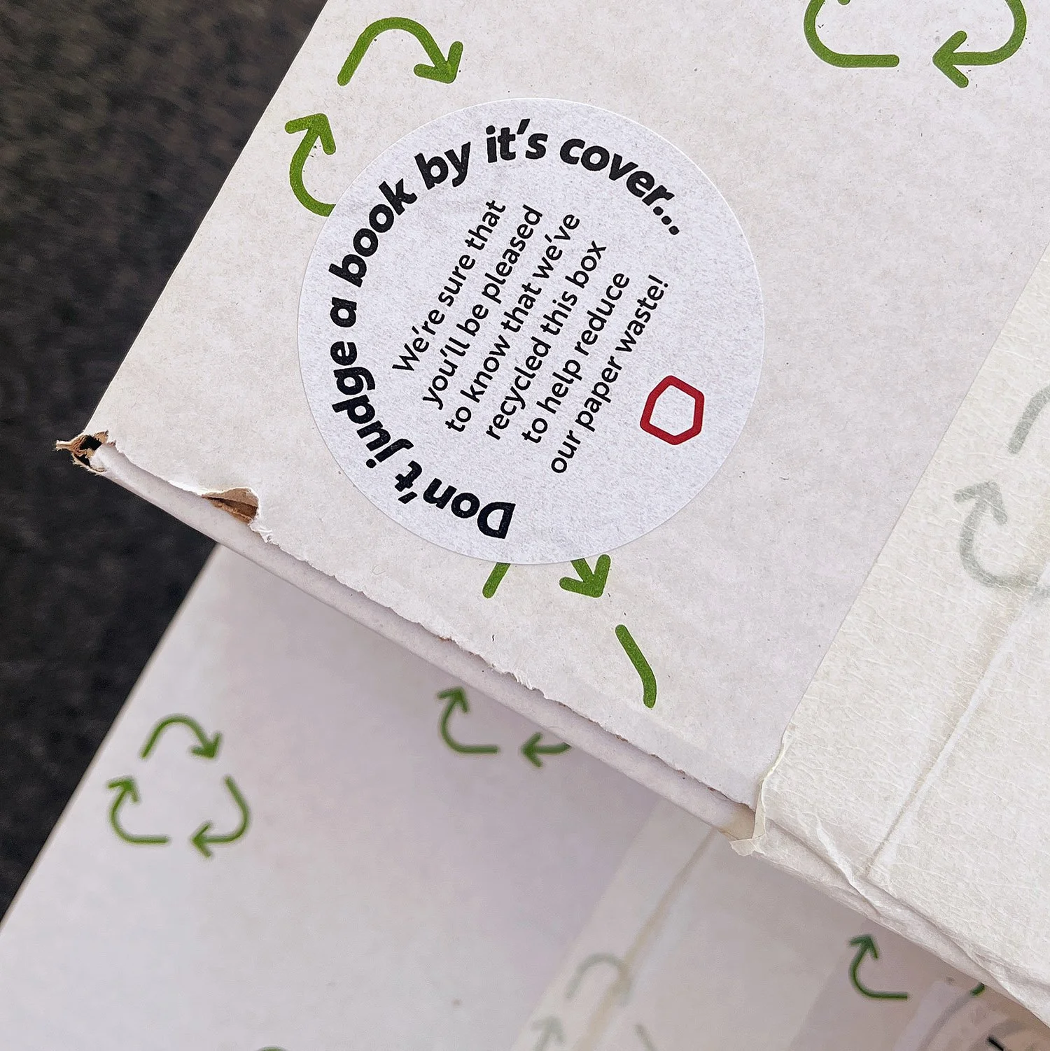 Close-up of a white cardboard box with a circular label indicating it helps reduce paper waste by recycling the box, with green recycling symbols and a small red outline of a shape on the label.