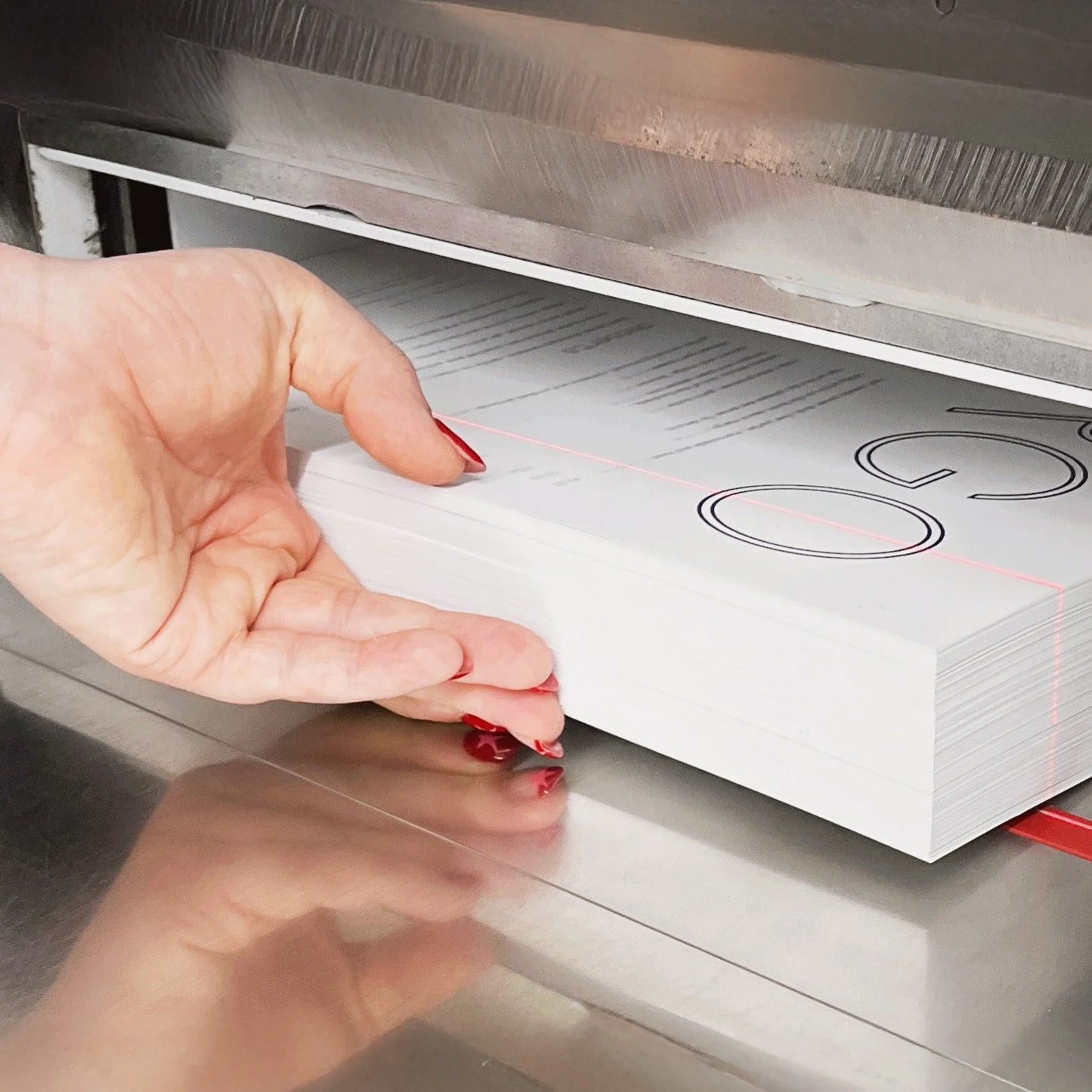 A person with red painted nails is placing a thick stack of printed sheets into a laser printer.