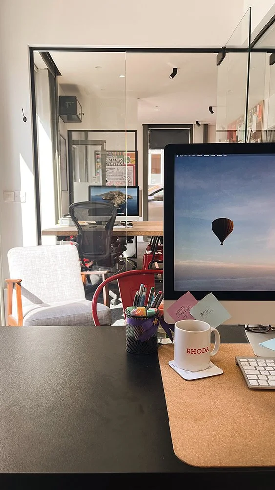 Office workspace with computer monitor displaying sky and hot air balloon, desk with mug, pens, and sticky notes, and additional chairs and computers in the background.