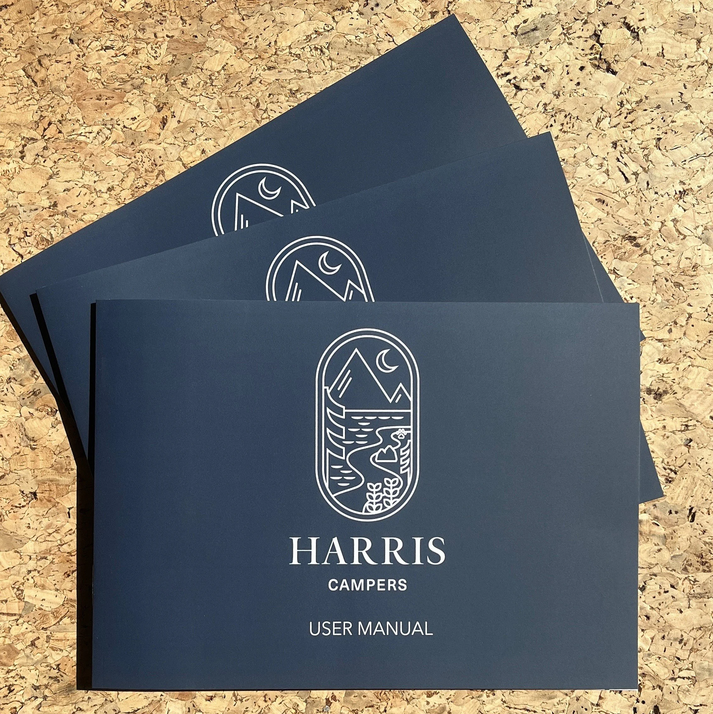 Three navy blue Harris Campers user manuals stacked on a cork surface with white line art of a mountain, a river, and a moon on the covers.