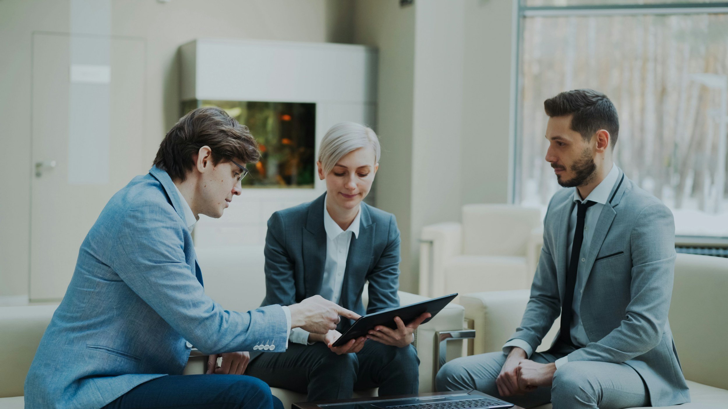 Three business professionals, two men and one woman, sit on a white sofa in a modern office or lobby, looking at a tablet device together.