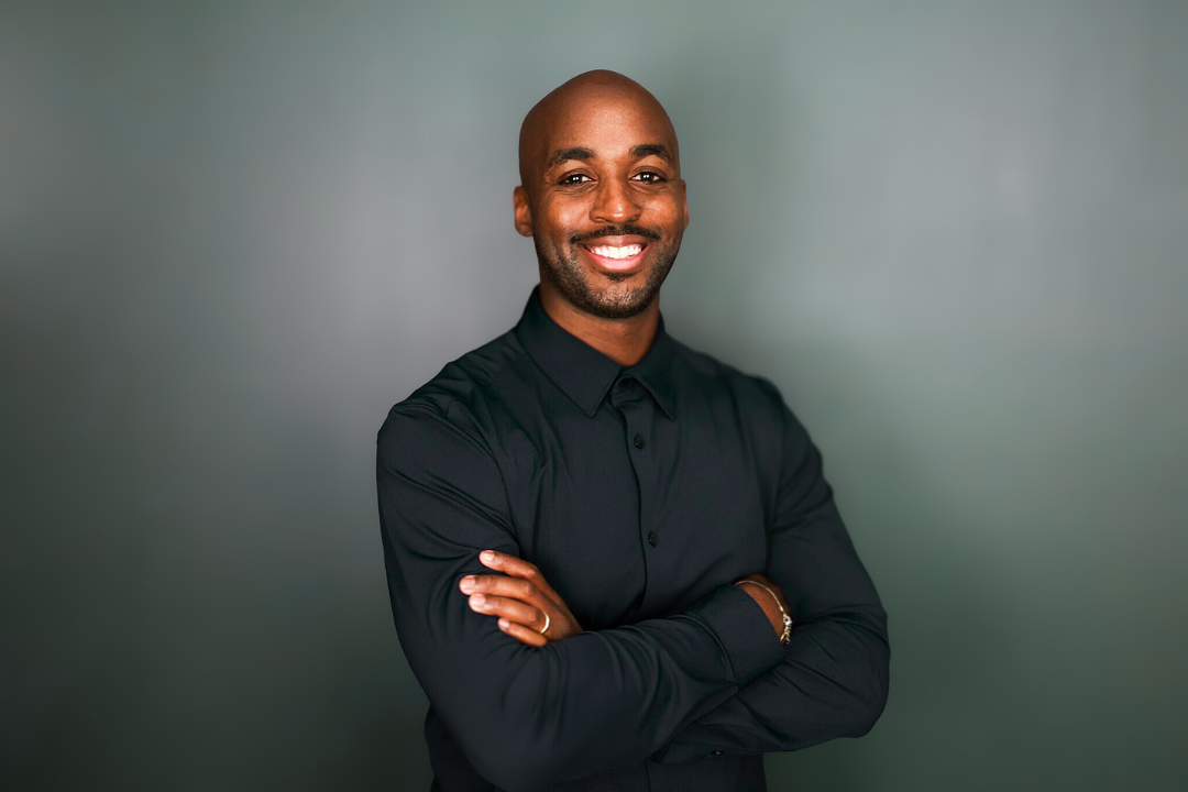 A smiling black man with a shaved head, wearing a black long-sleeve shirt, cross arms in front of a plain gray background.