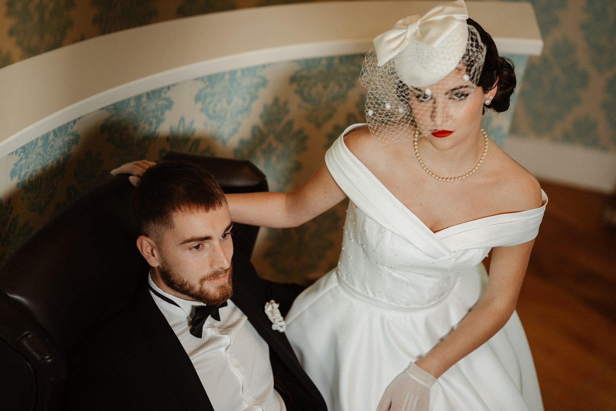 Mariée au maquillage vintage, lèvres rouges et voilette, assise près de son marié en smoking devant un papier peint à motifs bleus