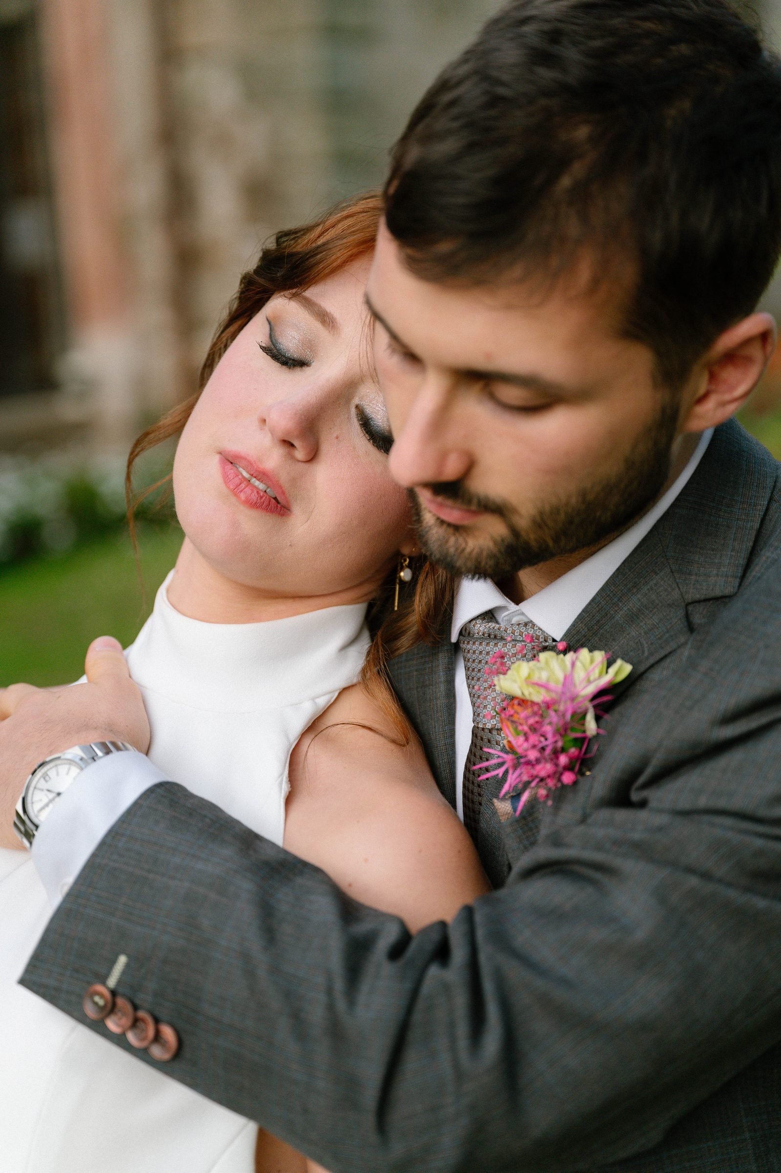 Mariée au maquillage lumineux et eye-liner graphique, la tête appuyée contre l'épaule de son marié, boutonnière fleurie rose