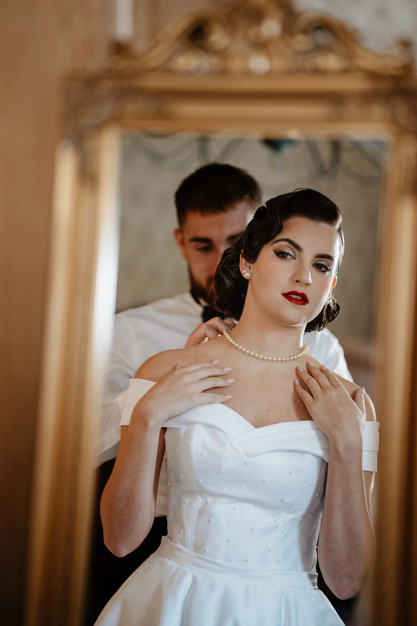 Mariée au maquillage rétro, lèvres rouges et collier de perles, devant un miroir doré où se reflète son marié