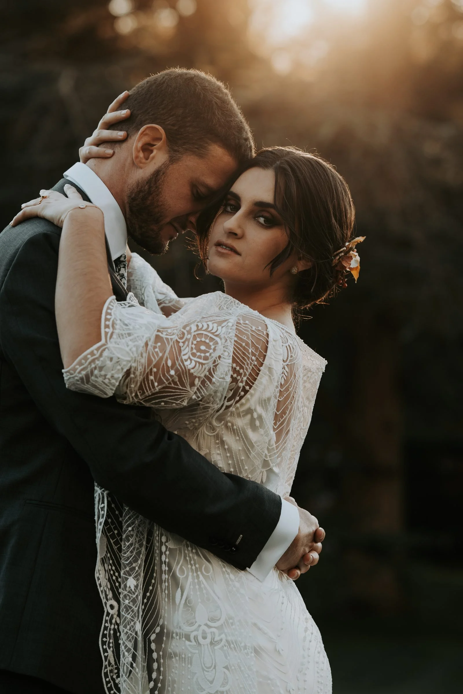 Mariée au regard intense, maquillage smoky et robe en dentelle brodée, enlacée par son marié dans une lumière dorée