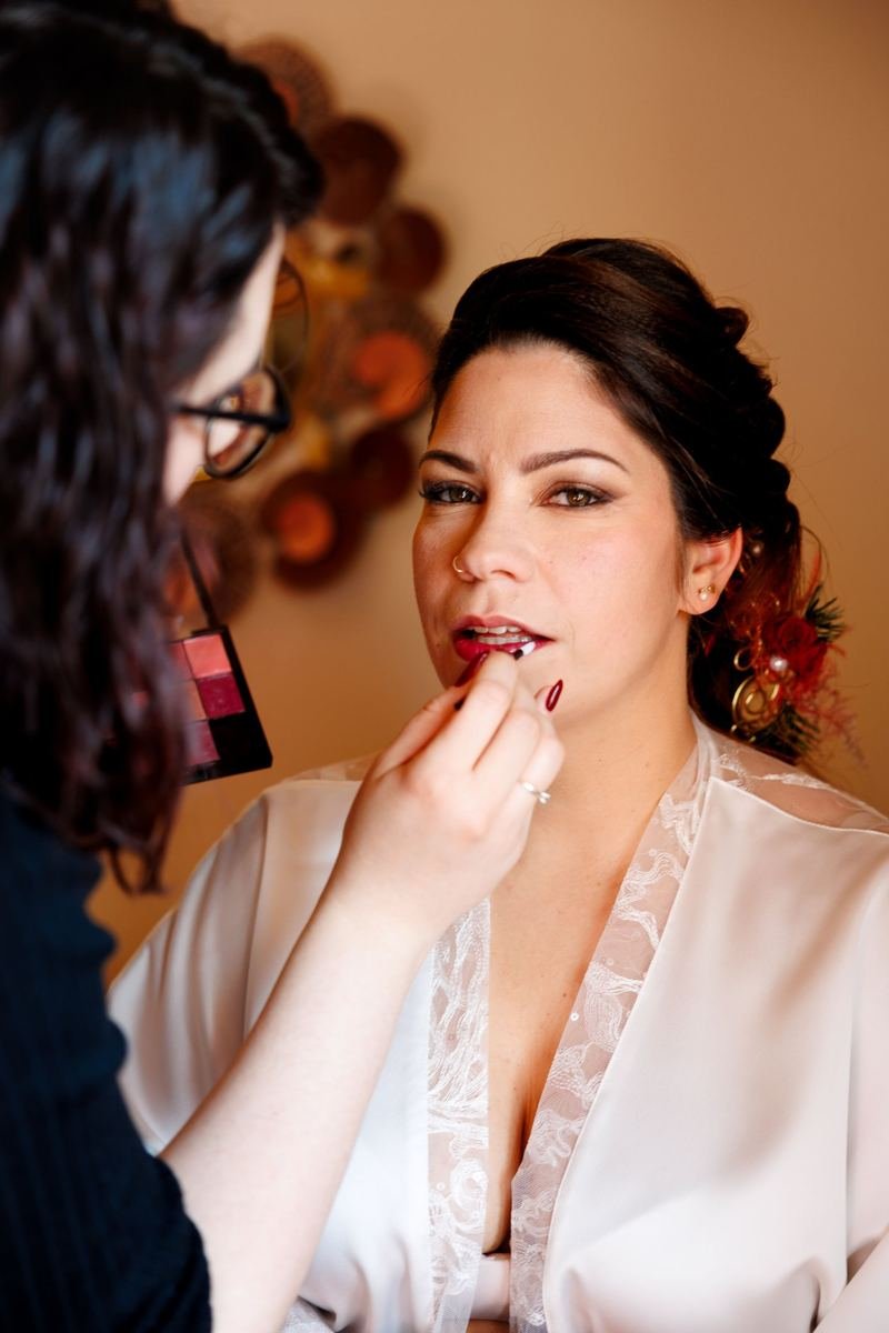 Préparatifs de mariage : mariée en peignoir de dentelle recevant le rouge à lèvres, palette de maquillage au premier plan