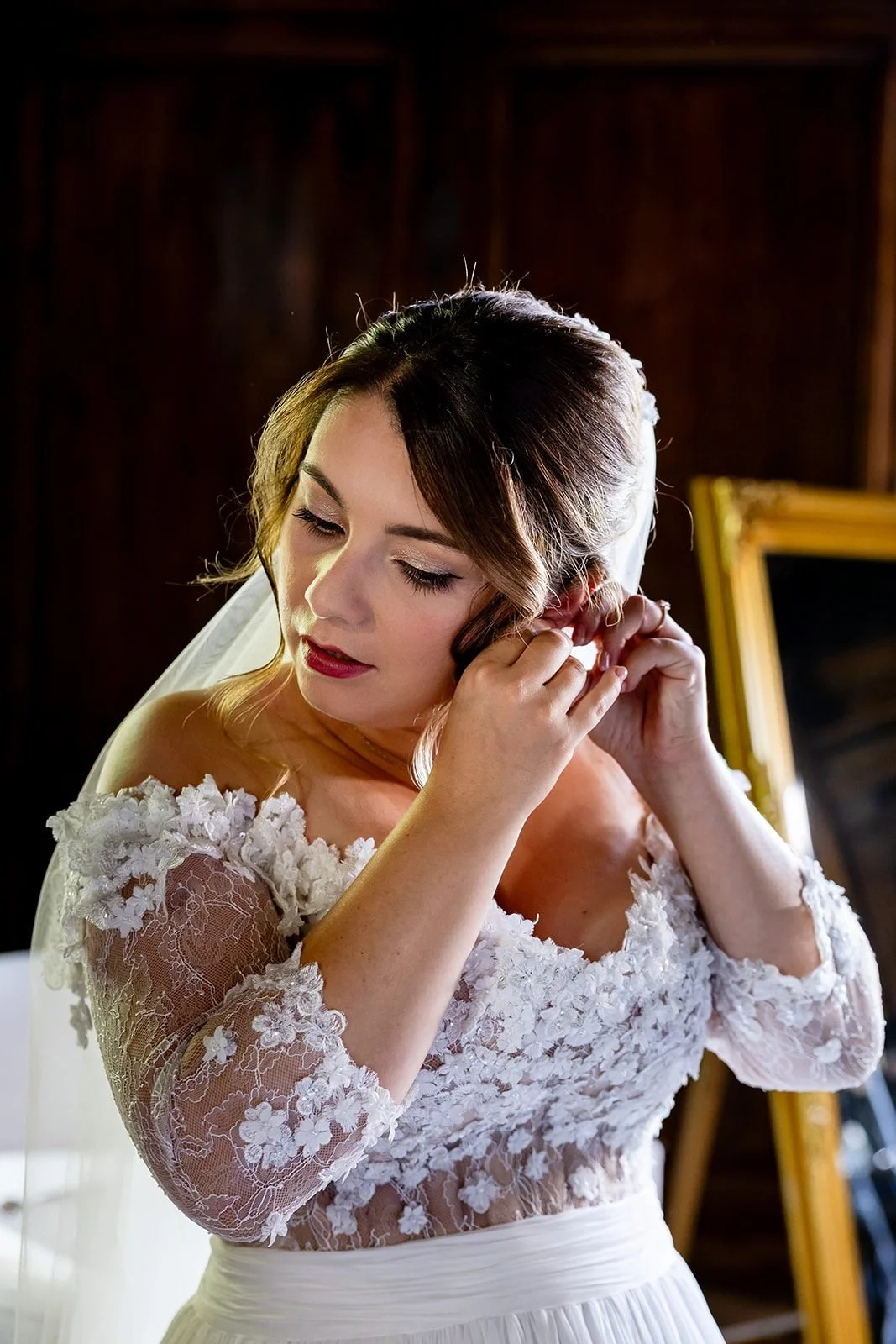 Candice ajustant sa boucle d'oreille lors des préparatifs : maquillage mariée élégant aux lèvres rouges, voile en dentelle