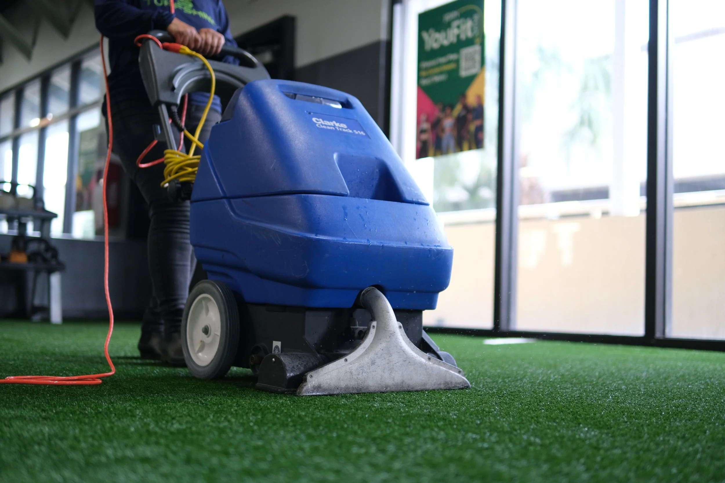 Close-up of a person operating a blue carpet cleaning machine on artificial green turf in a building with large glass windows.