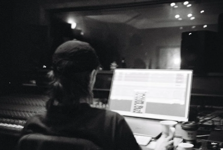 A person withlong hair in a ponytail working on a computer in a dimly lit studio or office, surrounded by coffee cups.