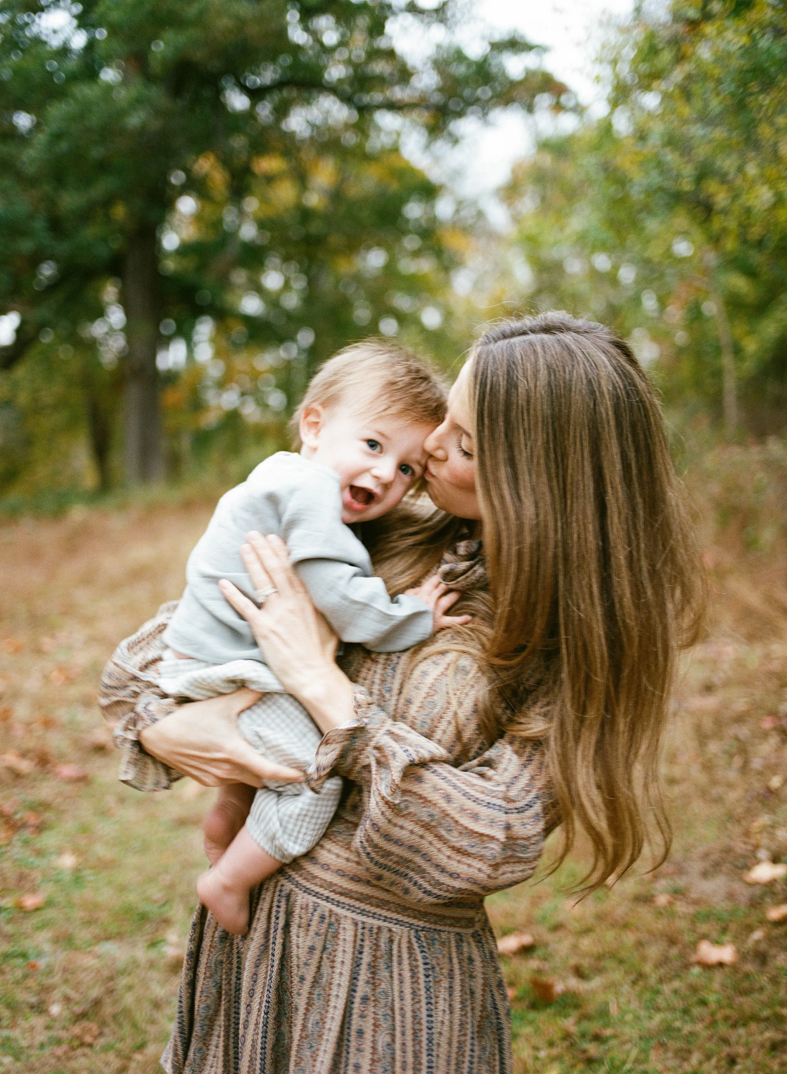 A woman holding a young boy outdoors in a wooded area during fall, with trees and leaves in the background.