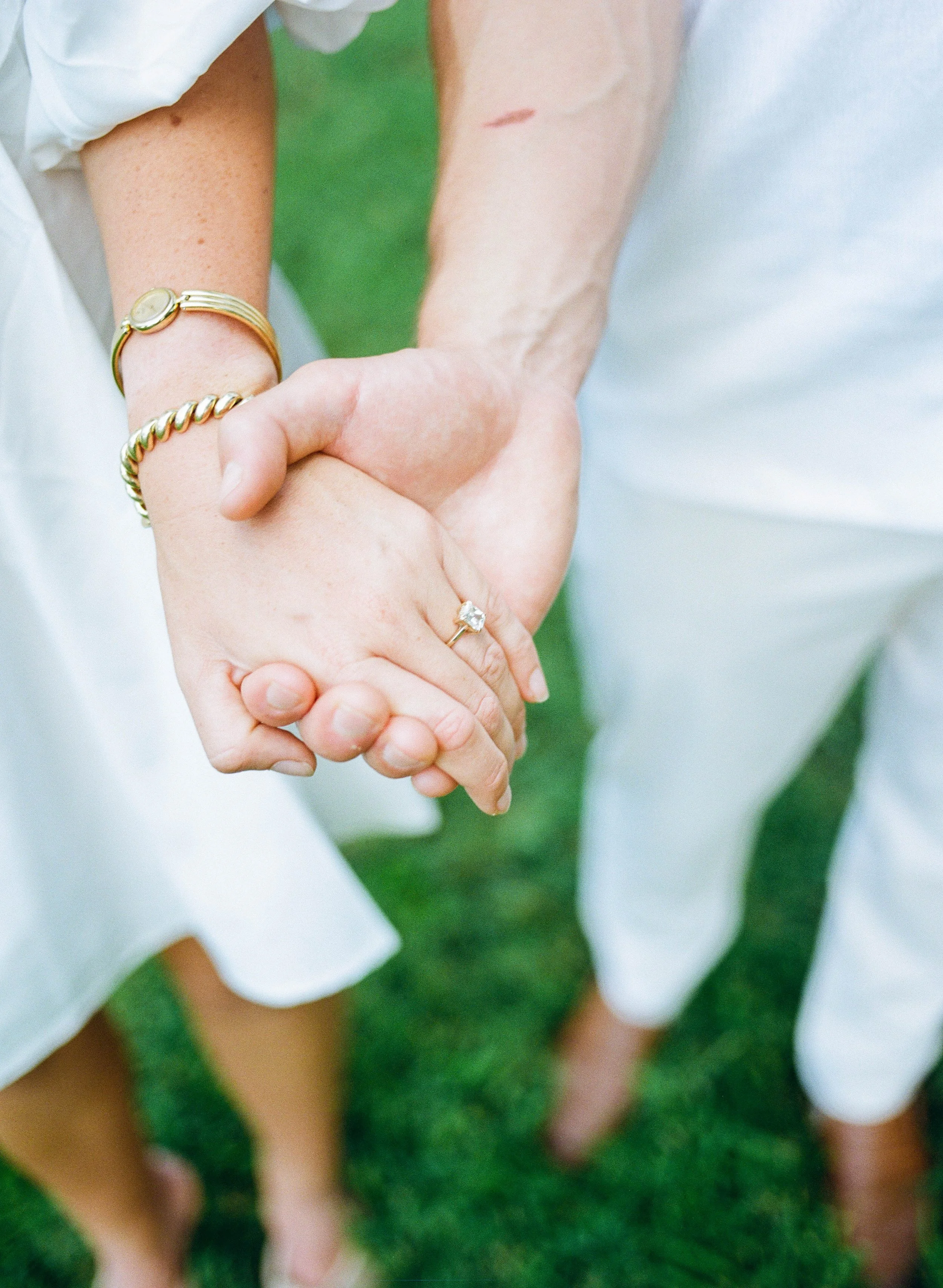 Close-up of a couple holding hands outdoors, with focus on their intertwined hands and wedding rings. The woman wears a gold bracelet, and the man has a small scratch on his arm. Both are dressed in white, with the background showing green grass.
