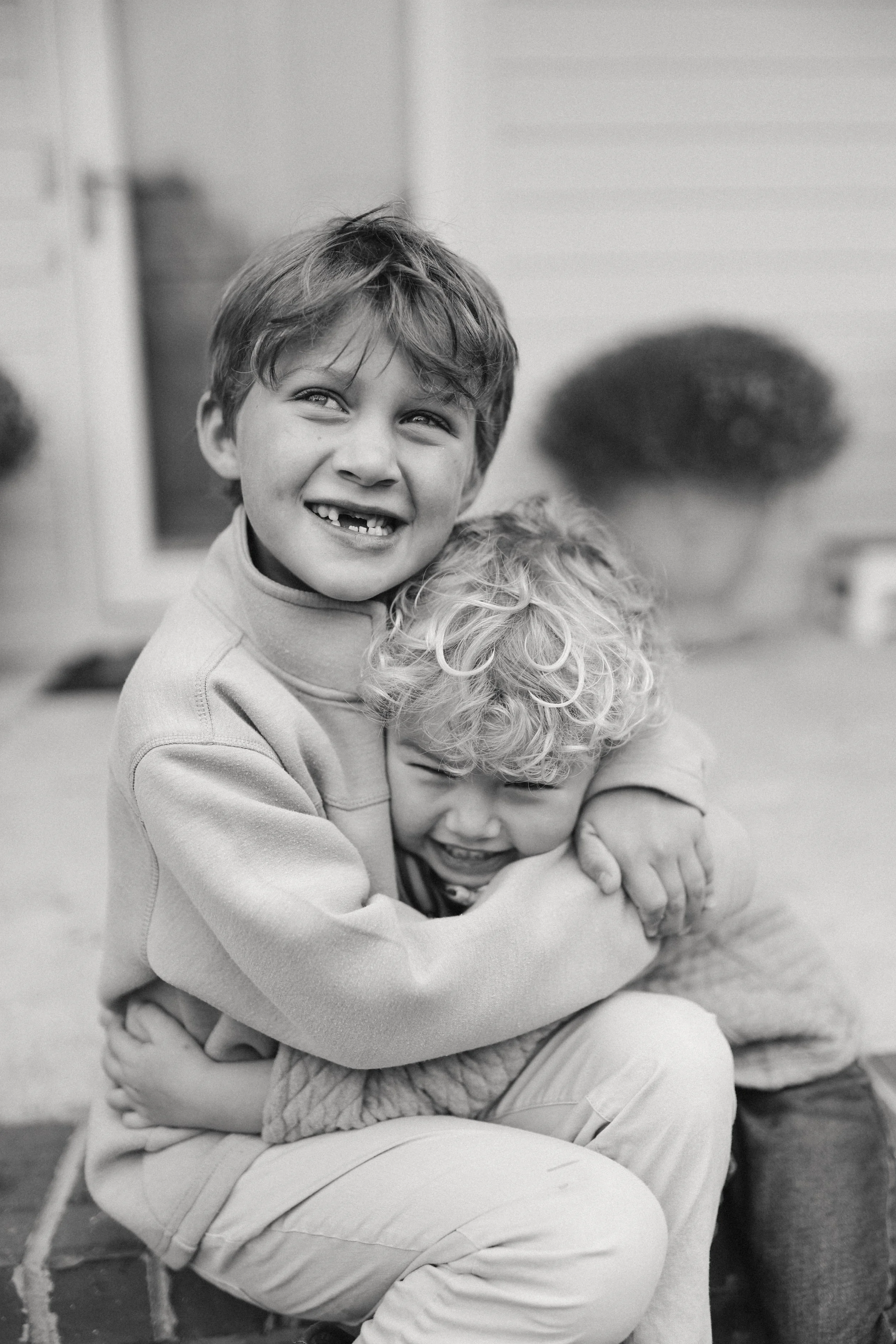 Two young boys hugging and smiling, sitting on a bench outdoors.