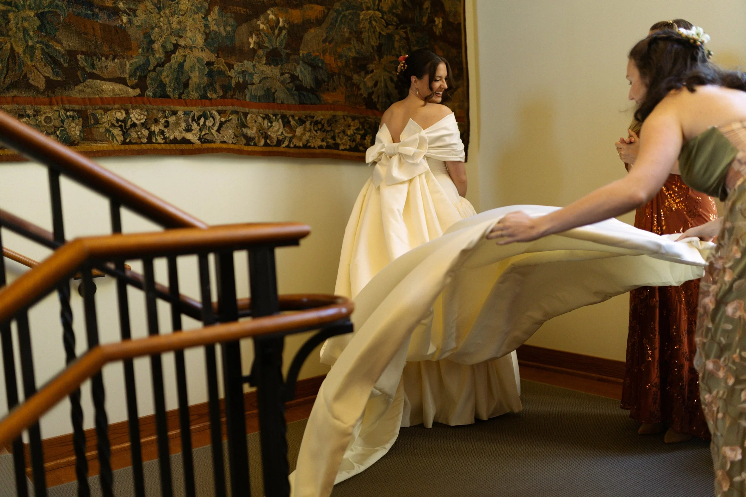 A woman in a yellow ball gown with a large bow on the back is being assisted with her dress by others as she prepares for a formal event.