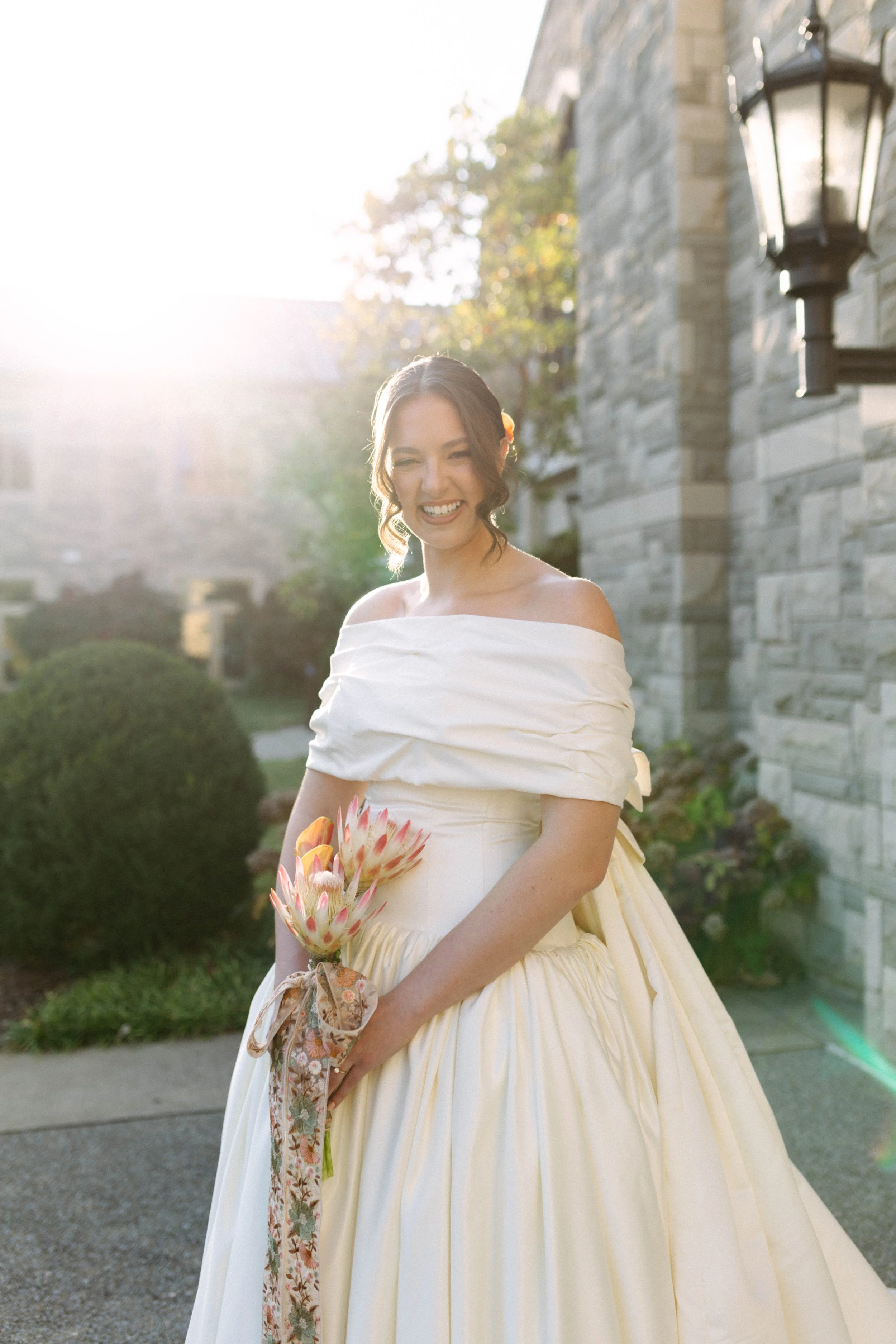 A woman in a white wedding dress holding flowers outdoors in front of a stone building with lanterns and greenery, with sunlight shining behind her.