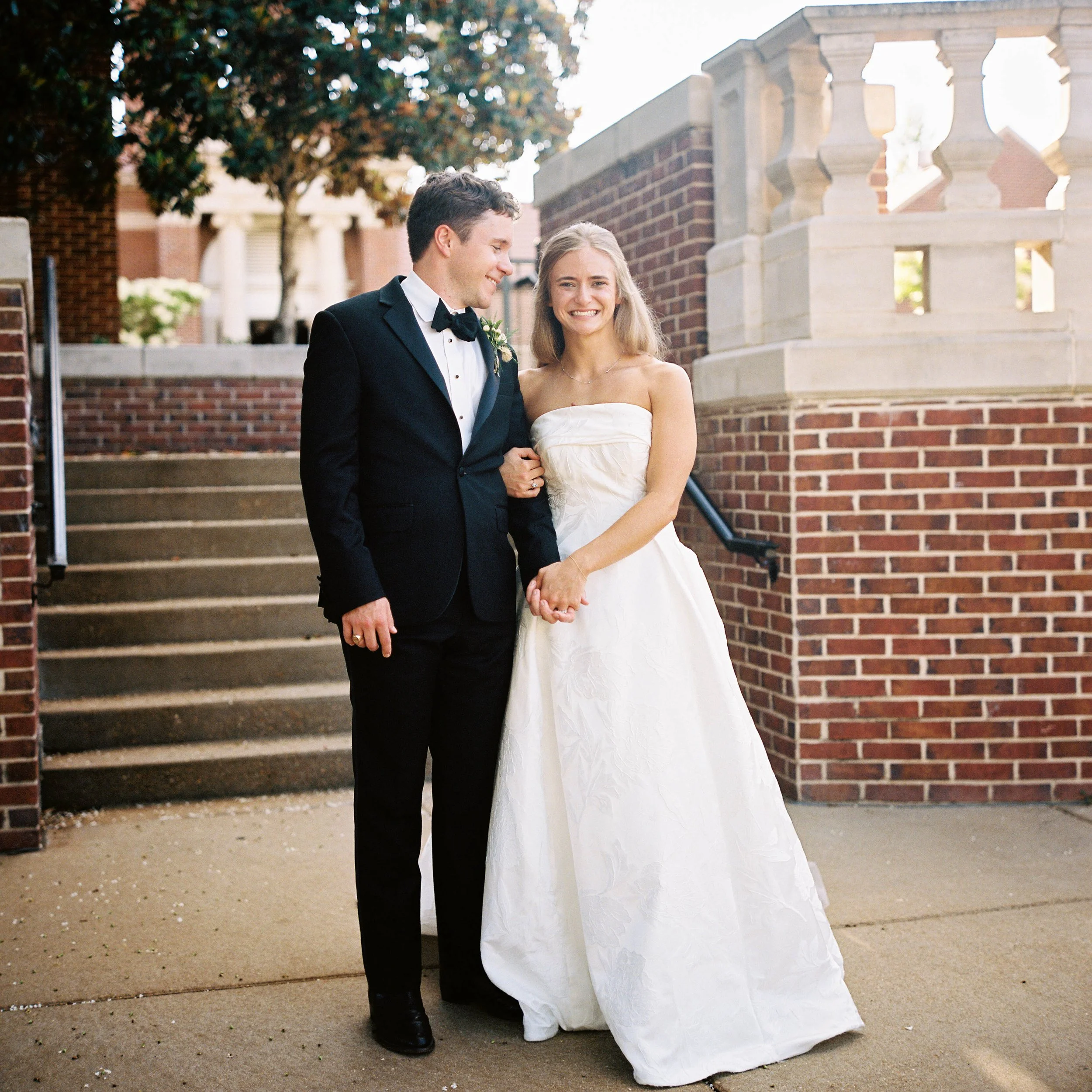 A smiling bride and groom standing outside on a brick staircase during their wedding, holding hands with the bride wearing a strapless white wedding dress and the groom in a black tuxedo with a bow tie.