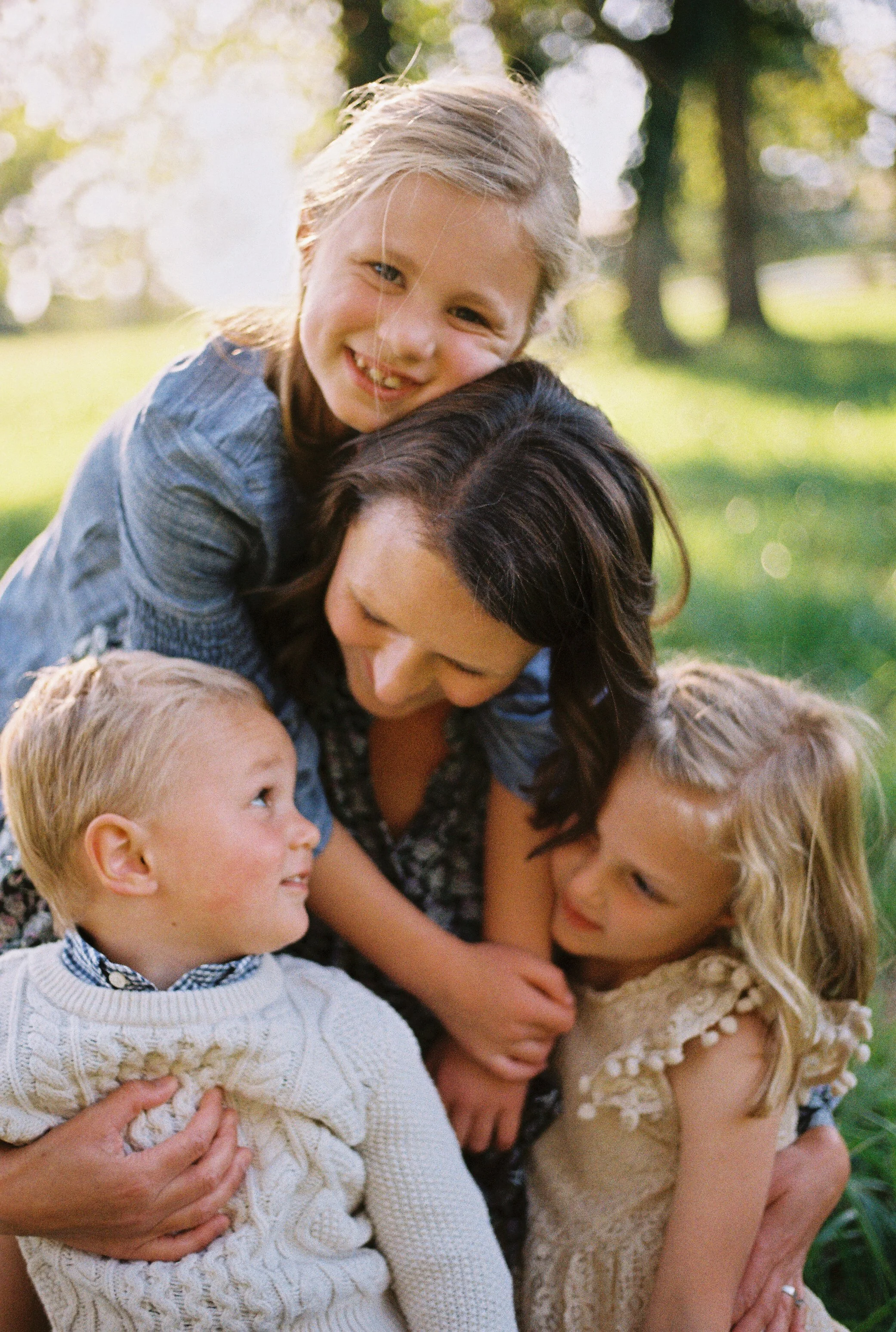 A woman and three children outdoors, hugging and smiling at each other in a park with trees and sunlight in the background.