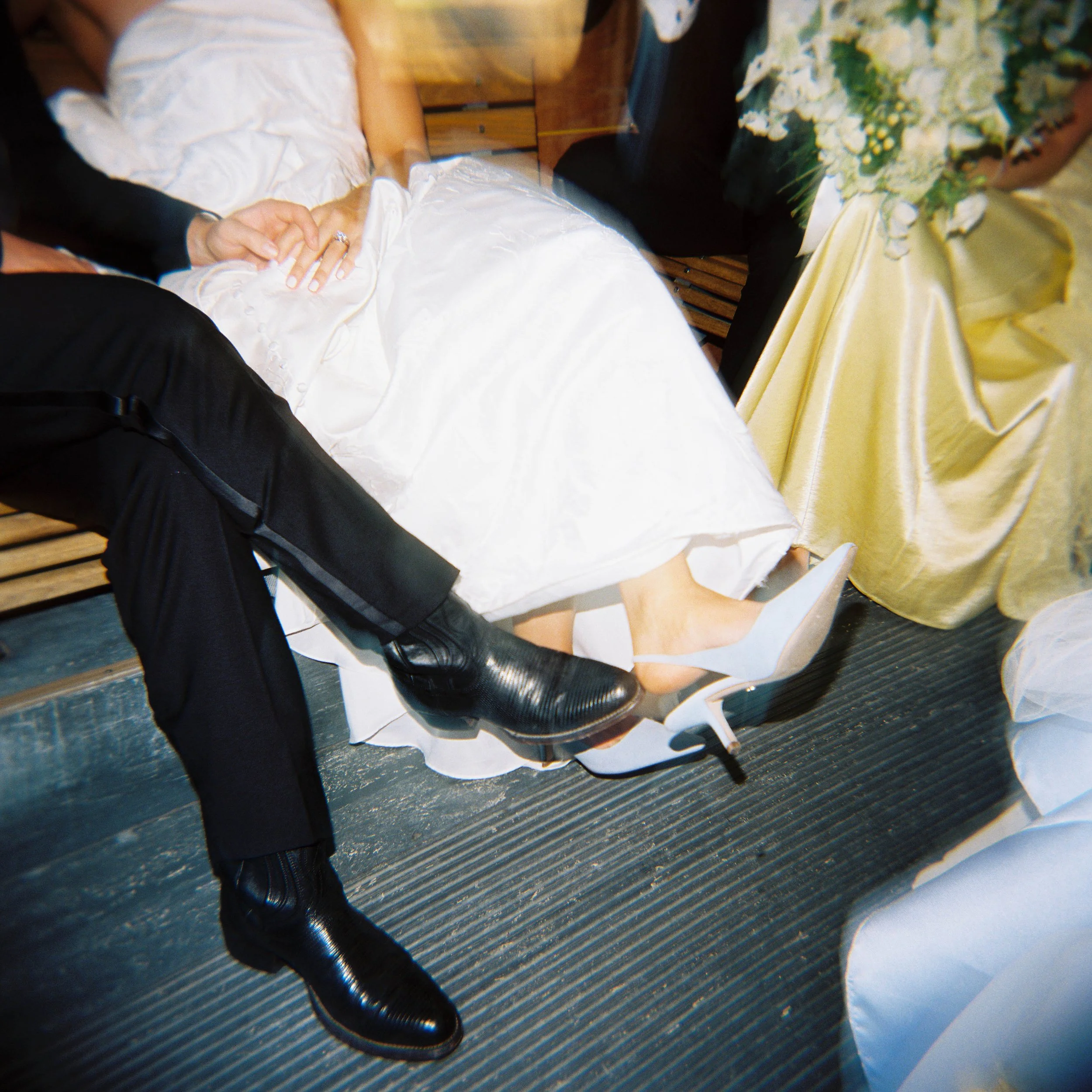 A close-up shot of a bride and groom holding hands during a wedding ceremony. The bride is wearing a white wedding dress with lace details, and the groom is dressed in a dark suit. They are sitting at a table with a yellow tablecloth, with a floral b