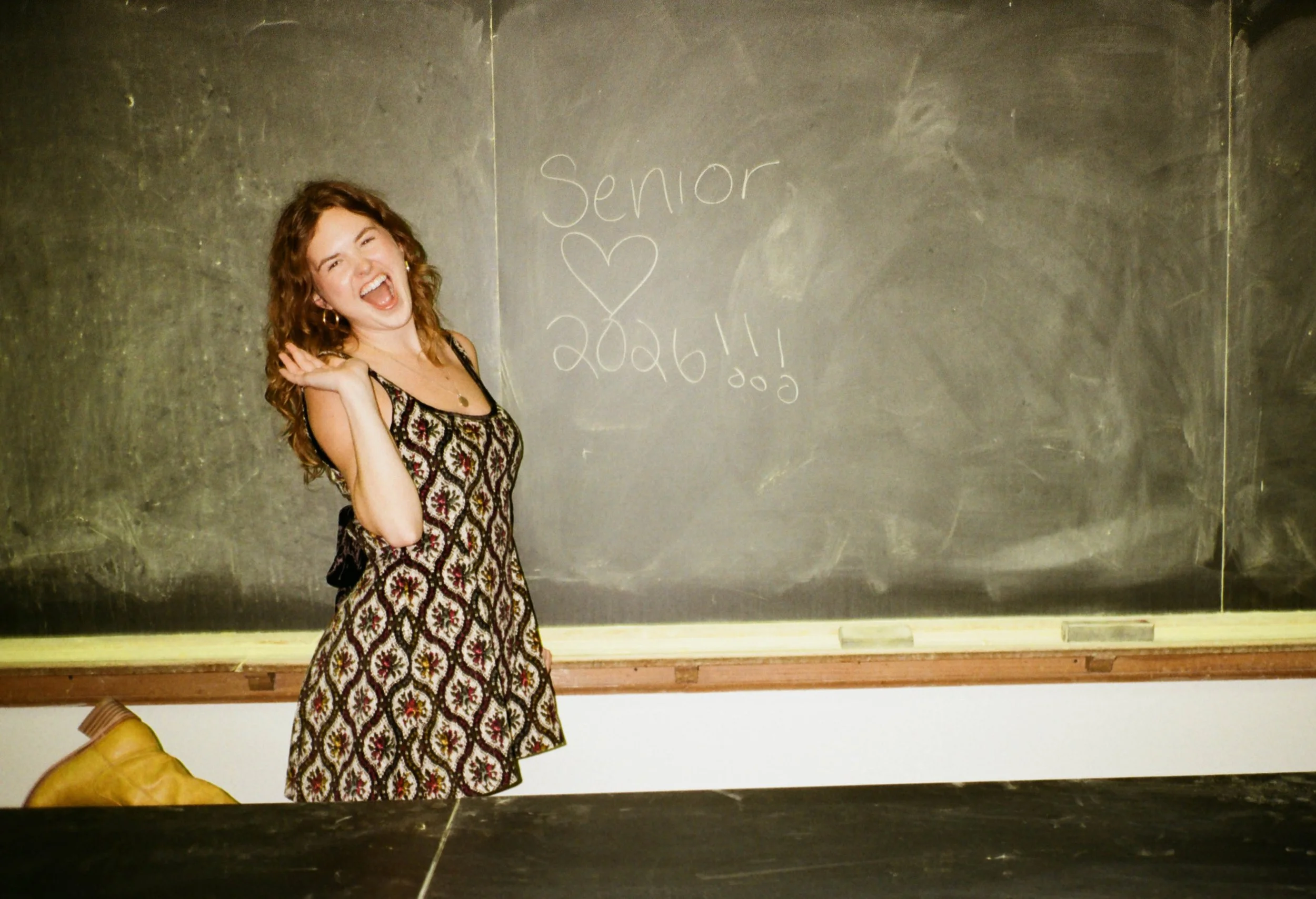 A young woman with curly hair in a patterned dress standing in front of a blackboard. She is smiling and waving, with the words 'Senior' and a heart drawn on the blackboard.