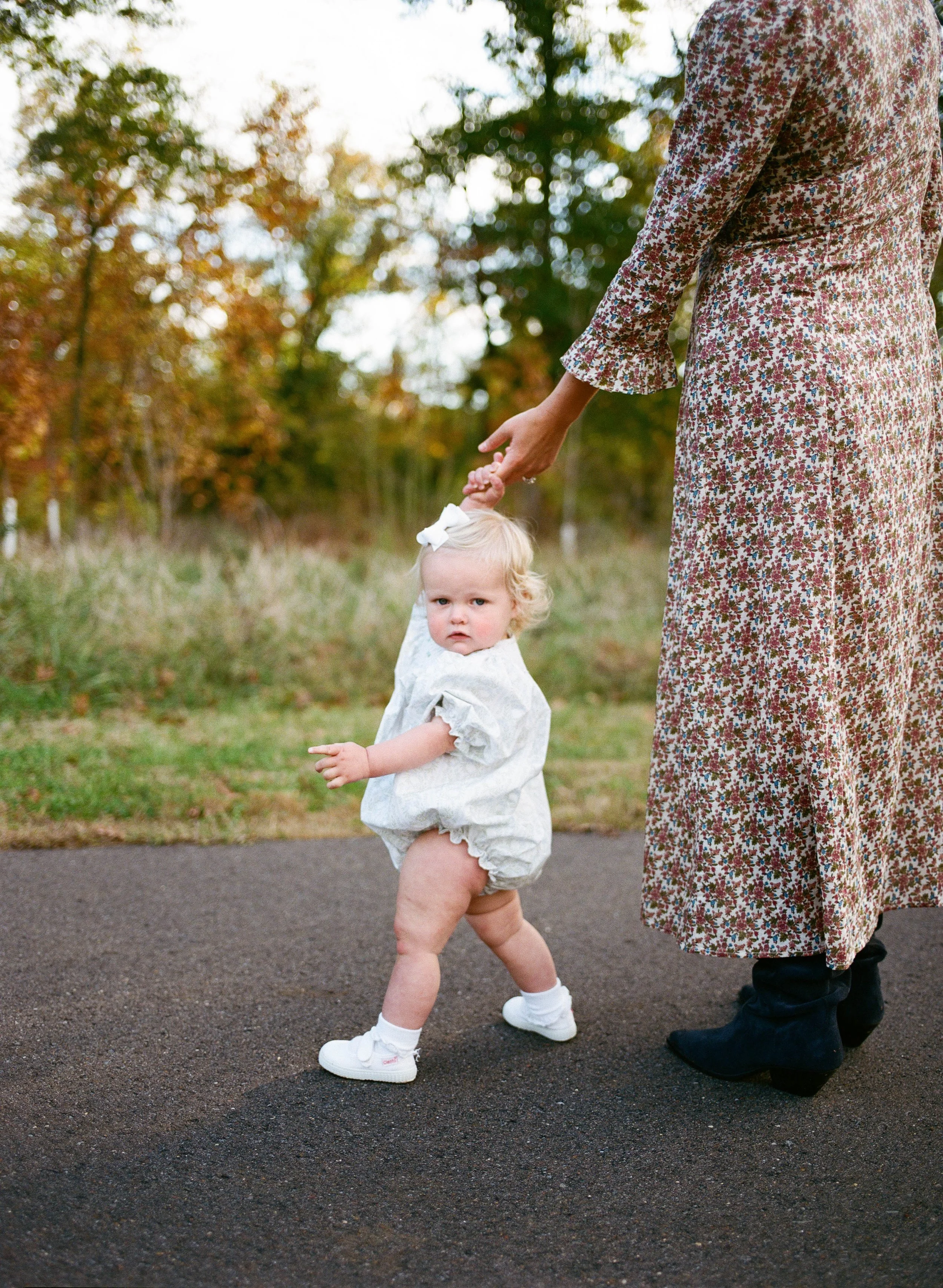 A toddler girl in a white dress with ruffled sleeves and a white bow in her blonde hair, walking while holding an adult woman’s hand. The woman is wearing a long, floral dress and black boots, and they are outdoors on a paved path surrounded by grass