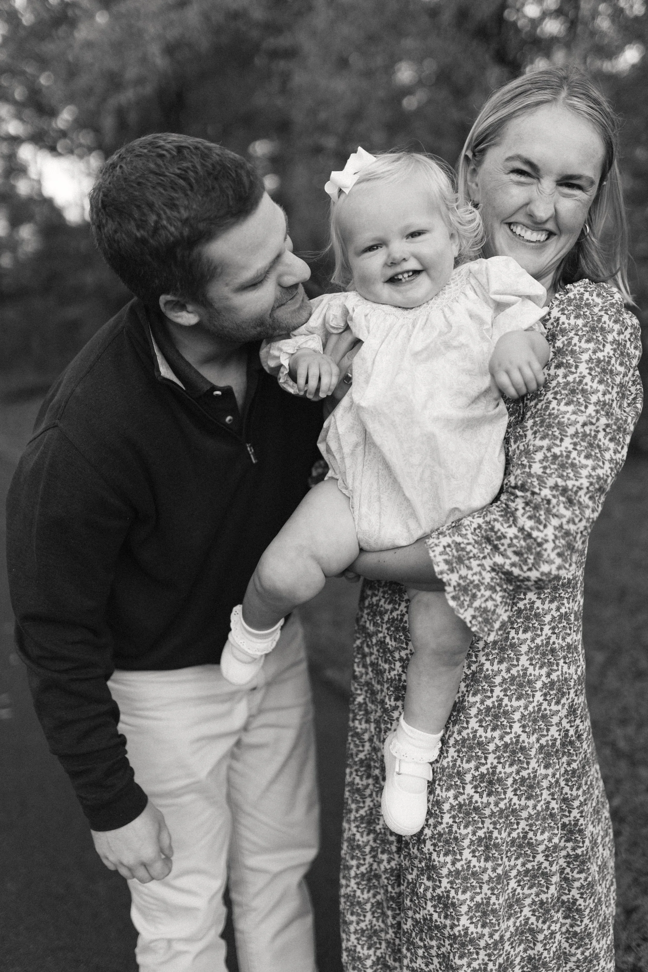 A smiling woman holding a young girl, with a man leaning in and kissing the girl on the cheek, outdoors in a park.