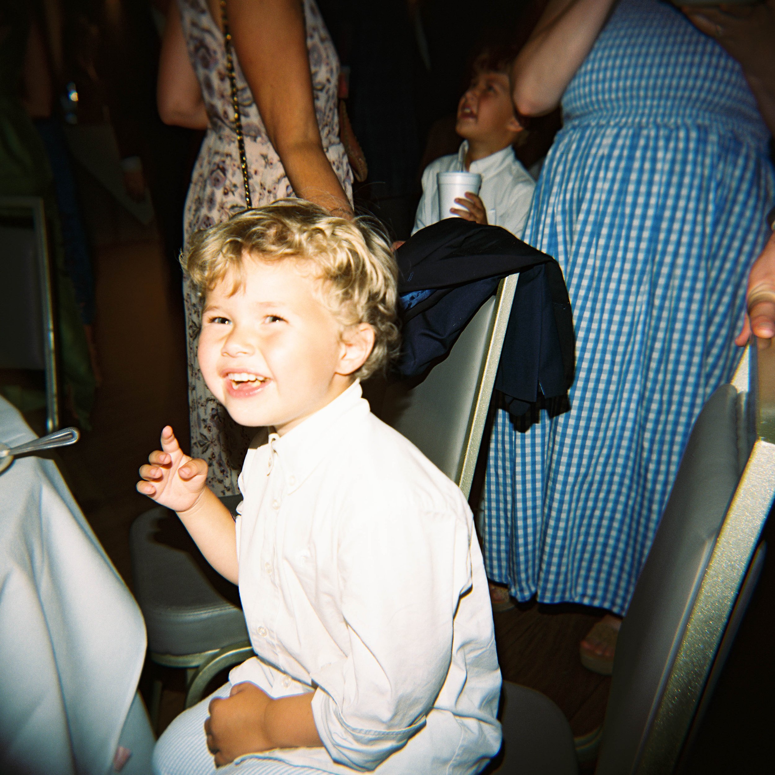 A young boy with curly blond hair smiling and laughing, sitting among adults in a crowded indoor space.