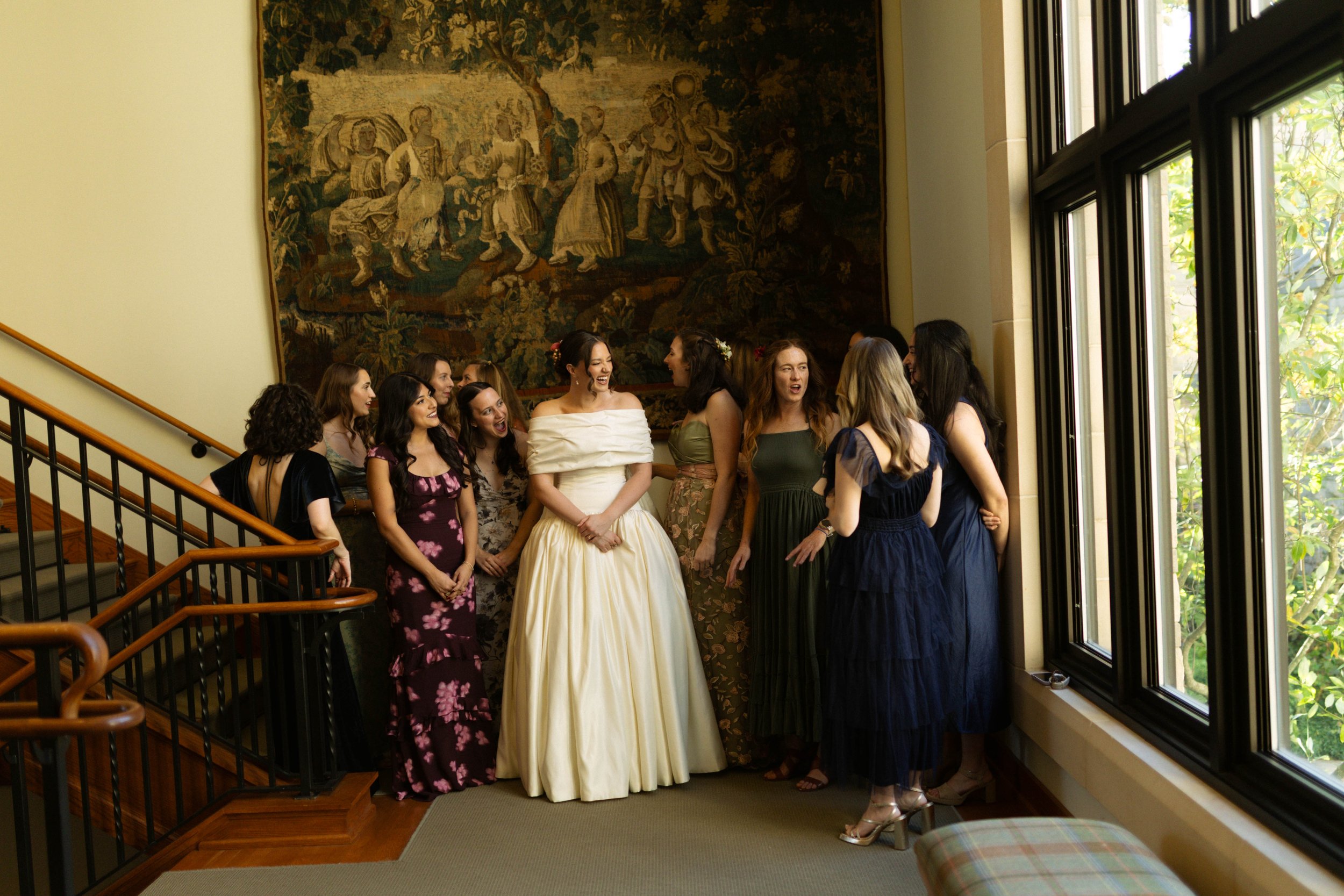 Group of women dressed in formal and evening attire standing in a hallway near a large window, engaged in conversation and laughter.