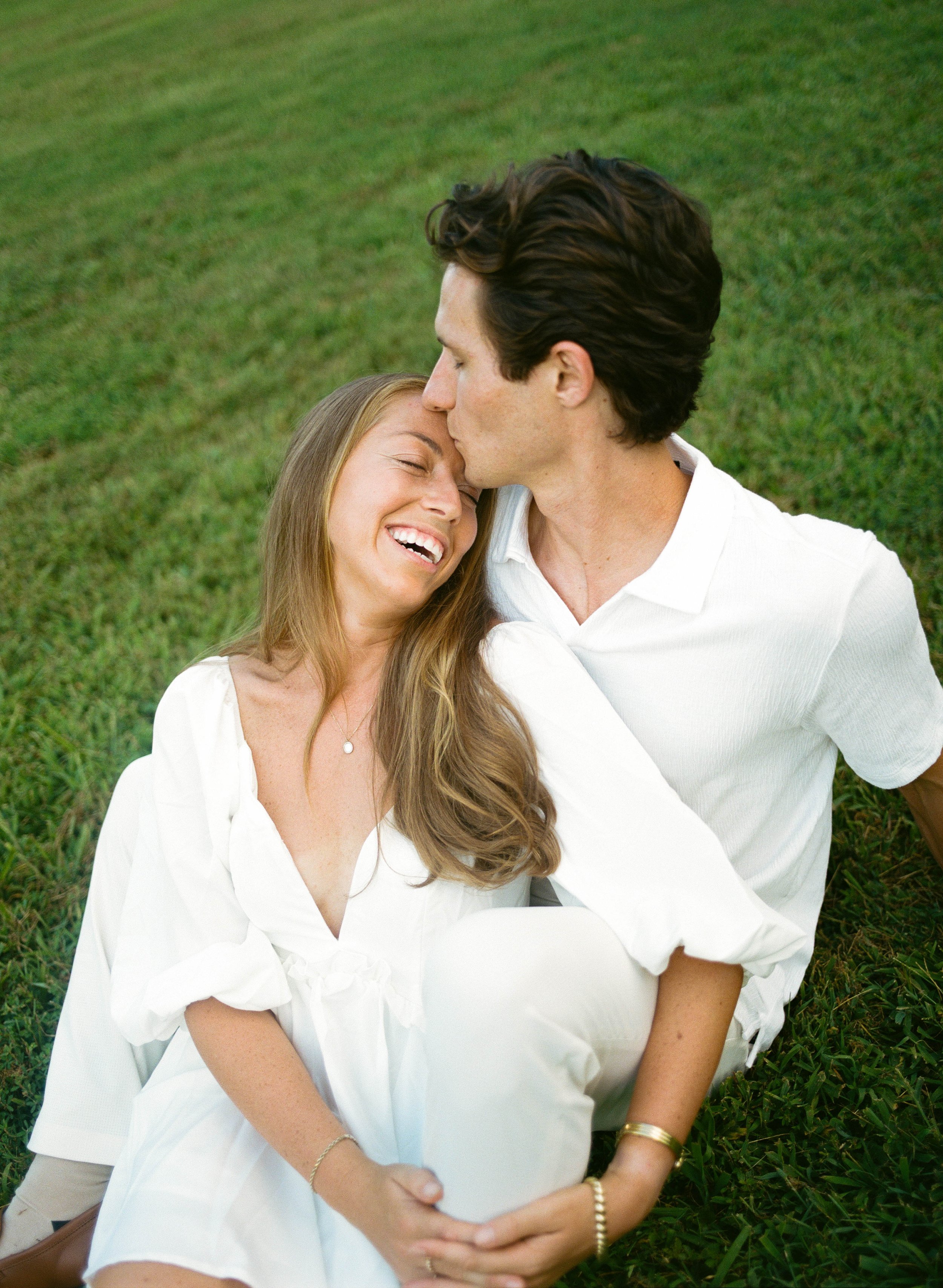 A couple sitting on grass, with the man kissing the woman's forehead. The woman is smiling with her eyes closed, wearing a white dress, and the man is dressed in white, leaning in for a kiss. Nashville film wedding photography, Southeast documentary 