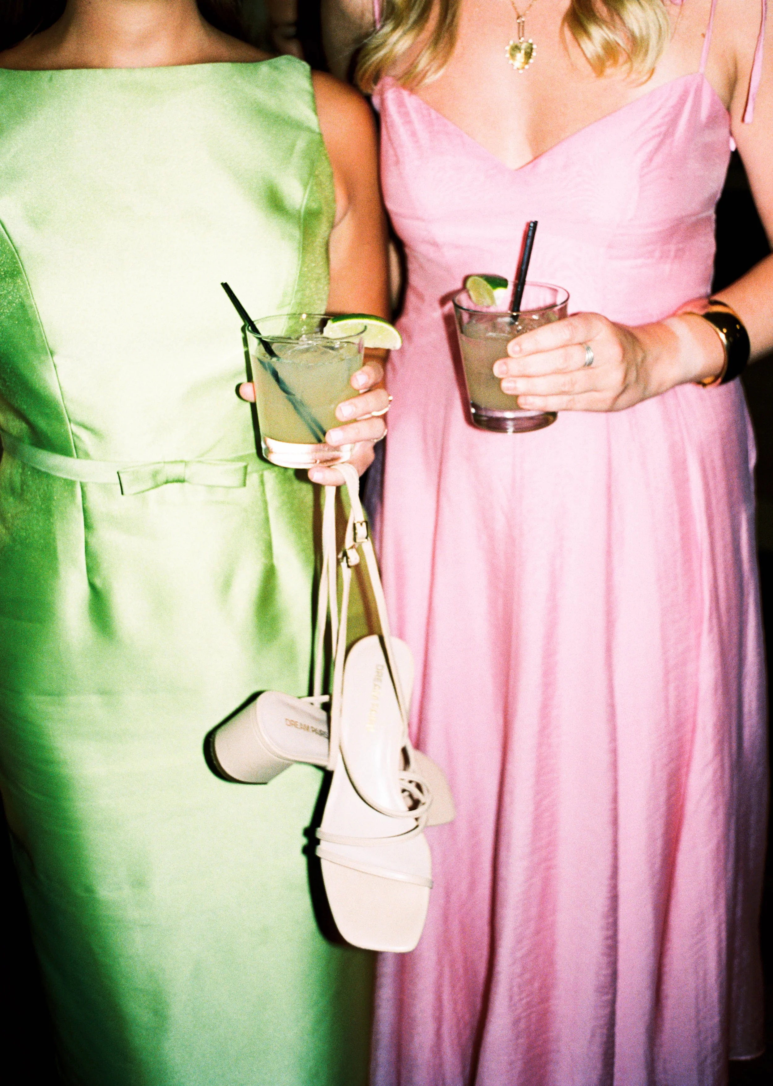Close-up of two women holding cocktails with lime wedges. One woman is wearing a bright green dress and white shoes, carrying a white handbag. The other woman is in a pink dress with a necklace and bracelets.