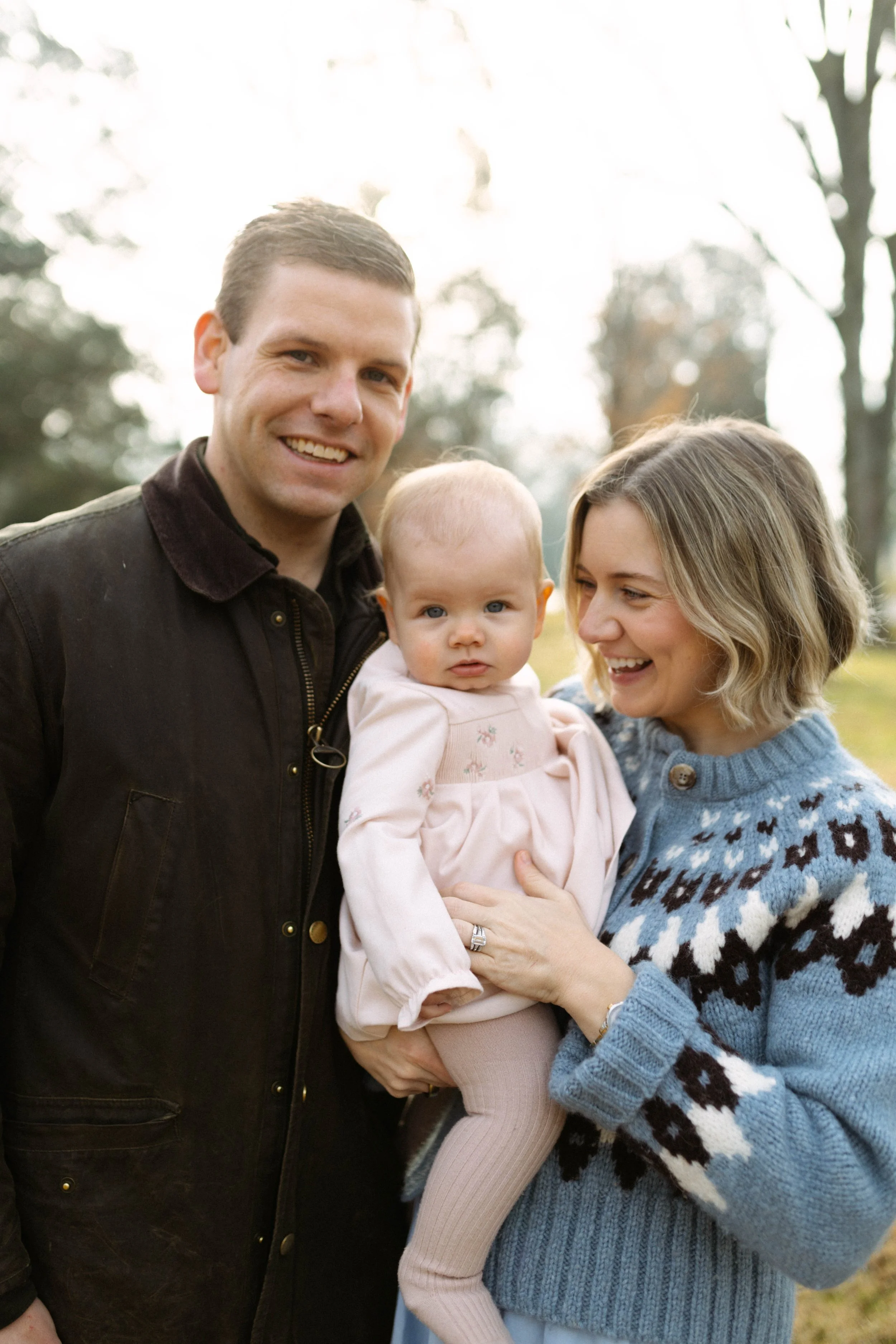 A smiling family of three outdoors, with a man, woman, and young girl with blue eyes and blonde hair, in a park with trees in the background.