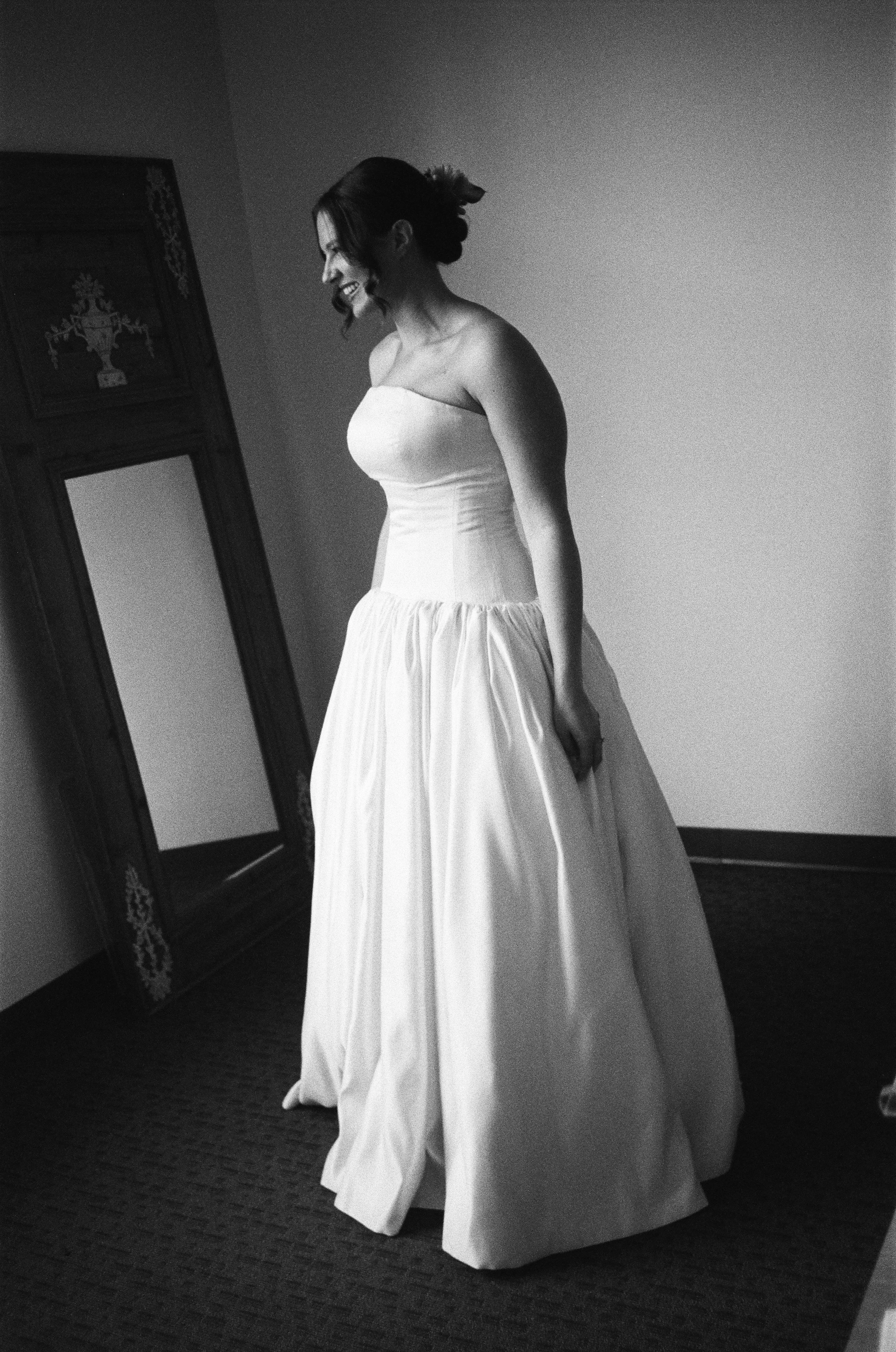 A woman in a strapless wedding dress, smiling and looking down, standing indoors in front of a mirror and plain wall.