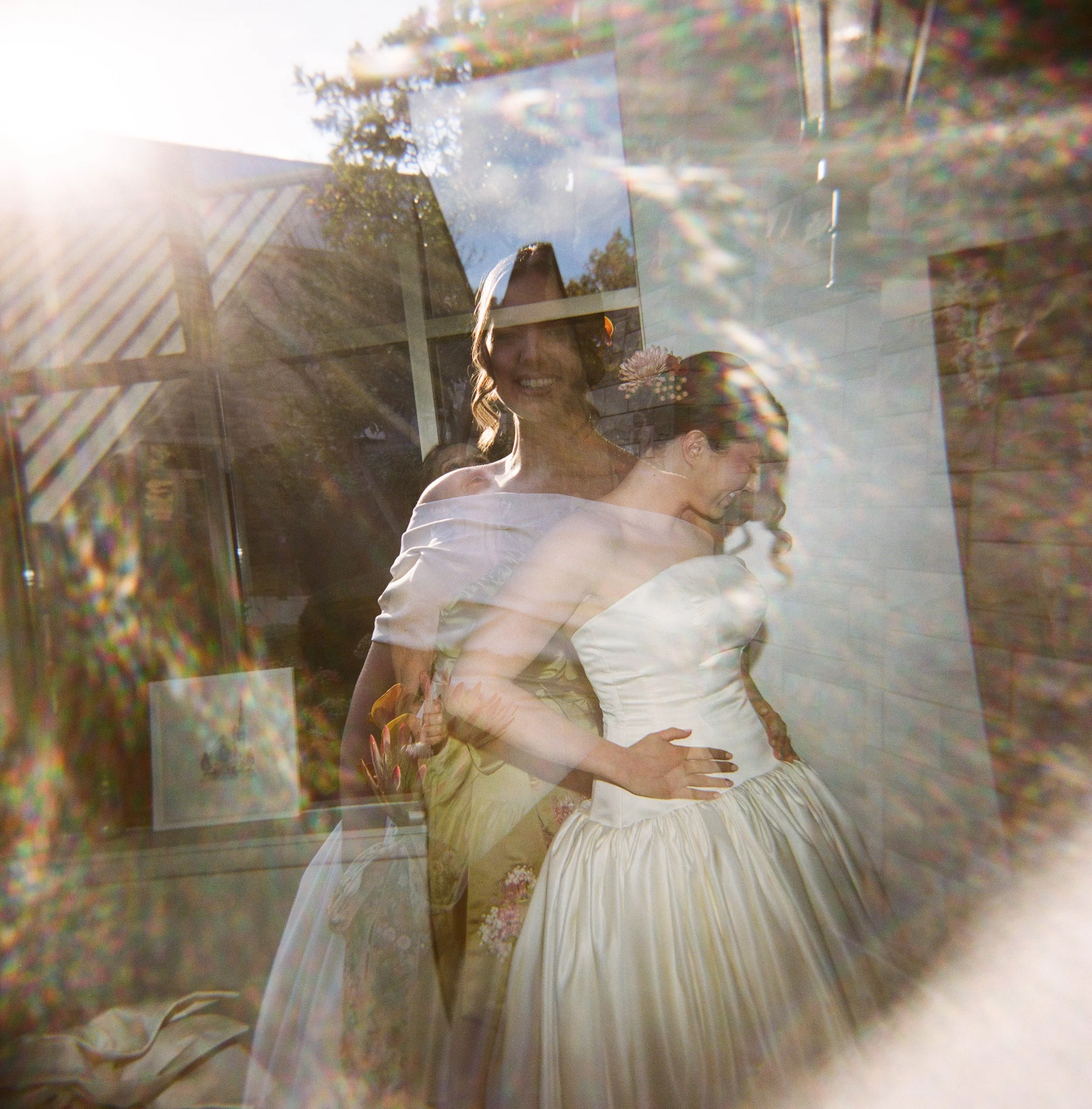 Two women in wedding dresses, one smiling, are captured through a glass window, creating a layered, artistic effect with sunlight and reflections.