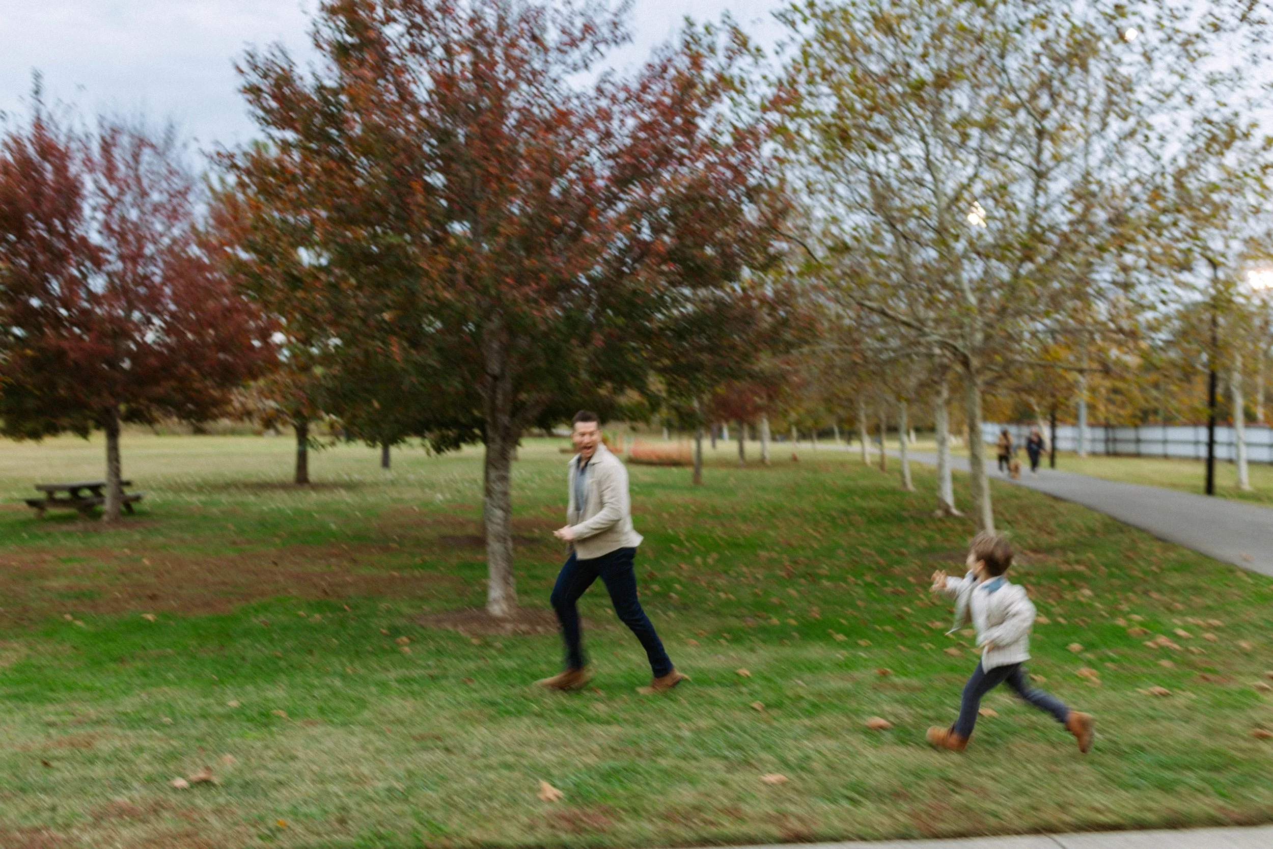 A father and son playing chase in a park during autumn, with colorful fall foliage and a walking path in the background.